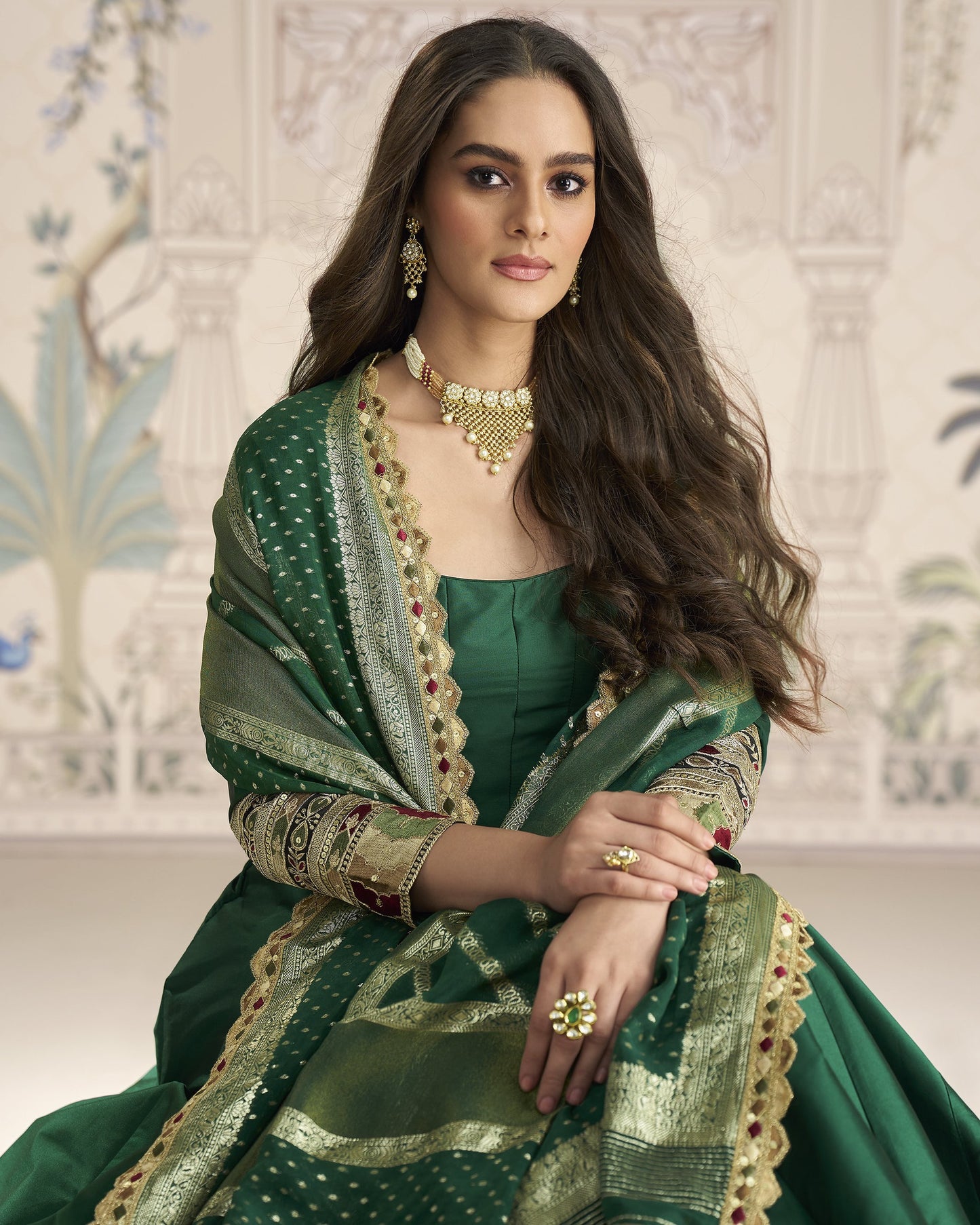 Woman in a green traditional outfit with jewelry against an ornate wall.