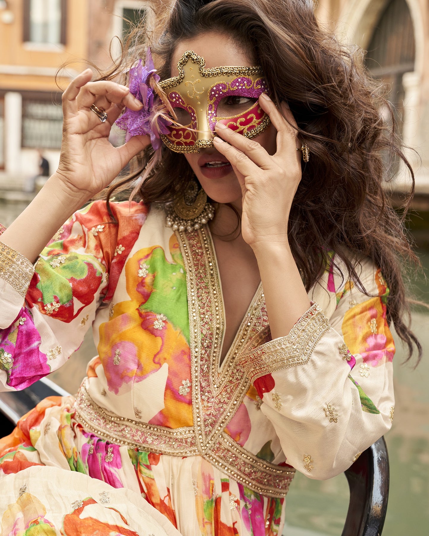 Woman in a colorful dress holding a decorative mask in front of her face, with a blurred background.