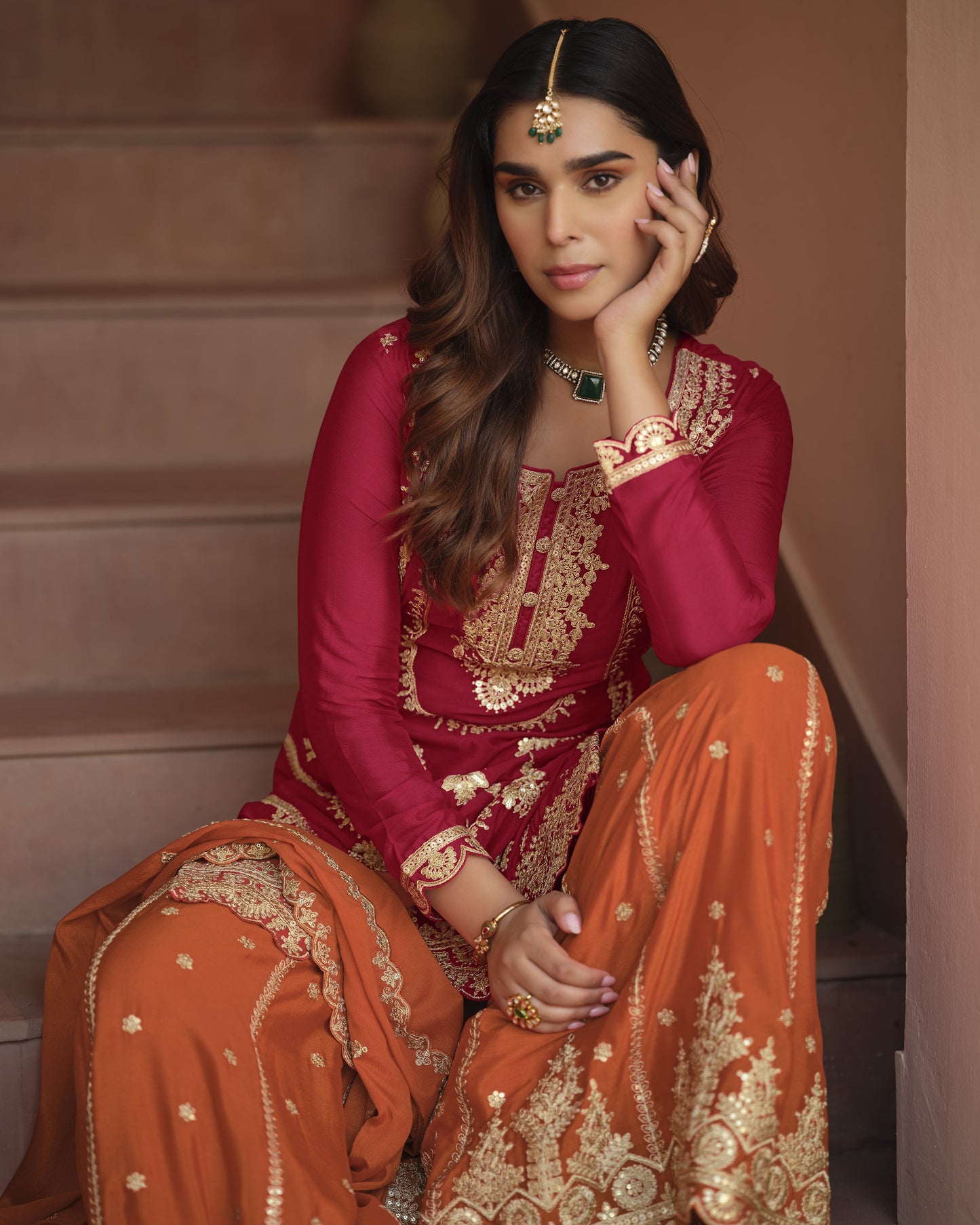 Woman in traditional pink and orange outfit with gold embroidery sitting on steps.