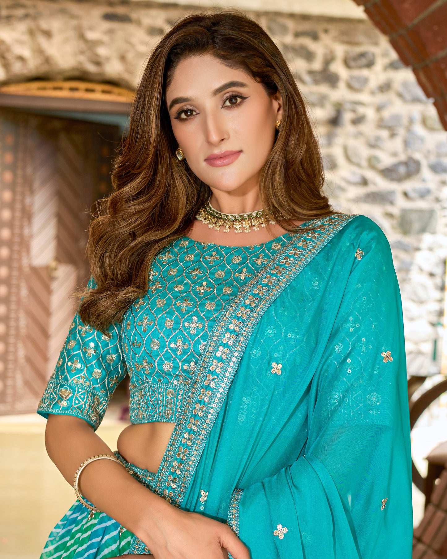 Woman wearing a teal saree with intricate patterns, standing against a stone wall background.