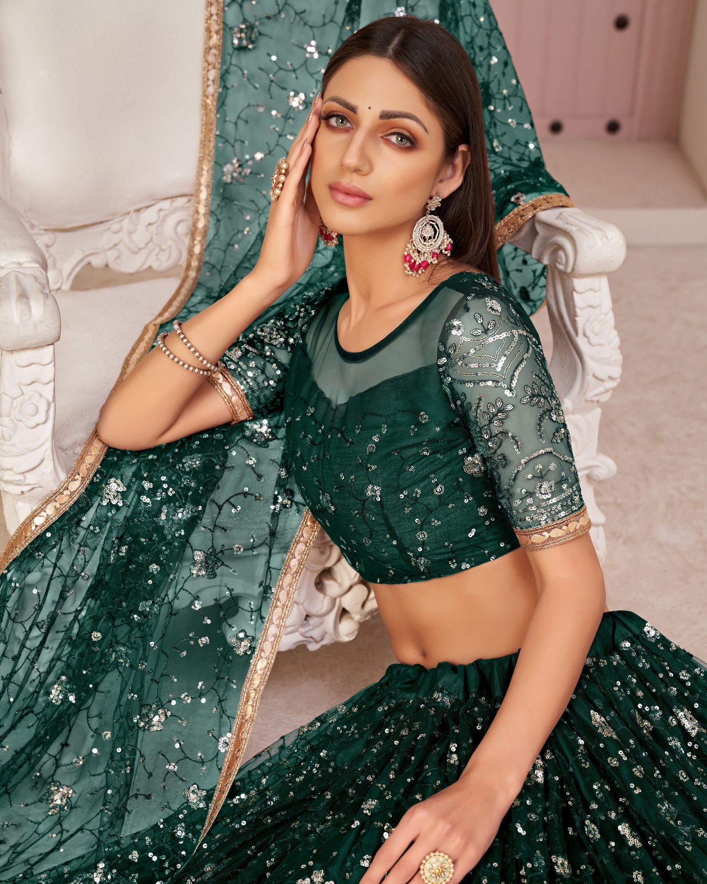 Woman in a green traditional outfit with sequin details, sitting on a decorative chair.