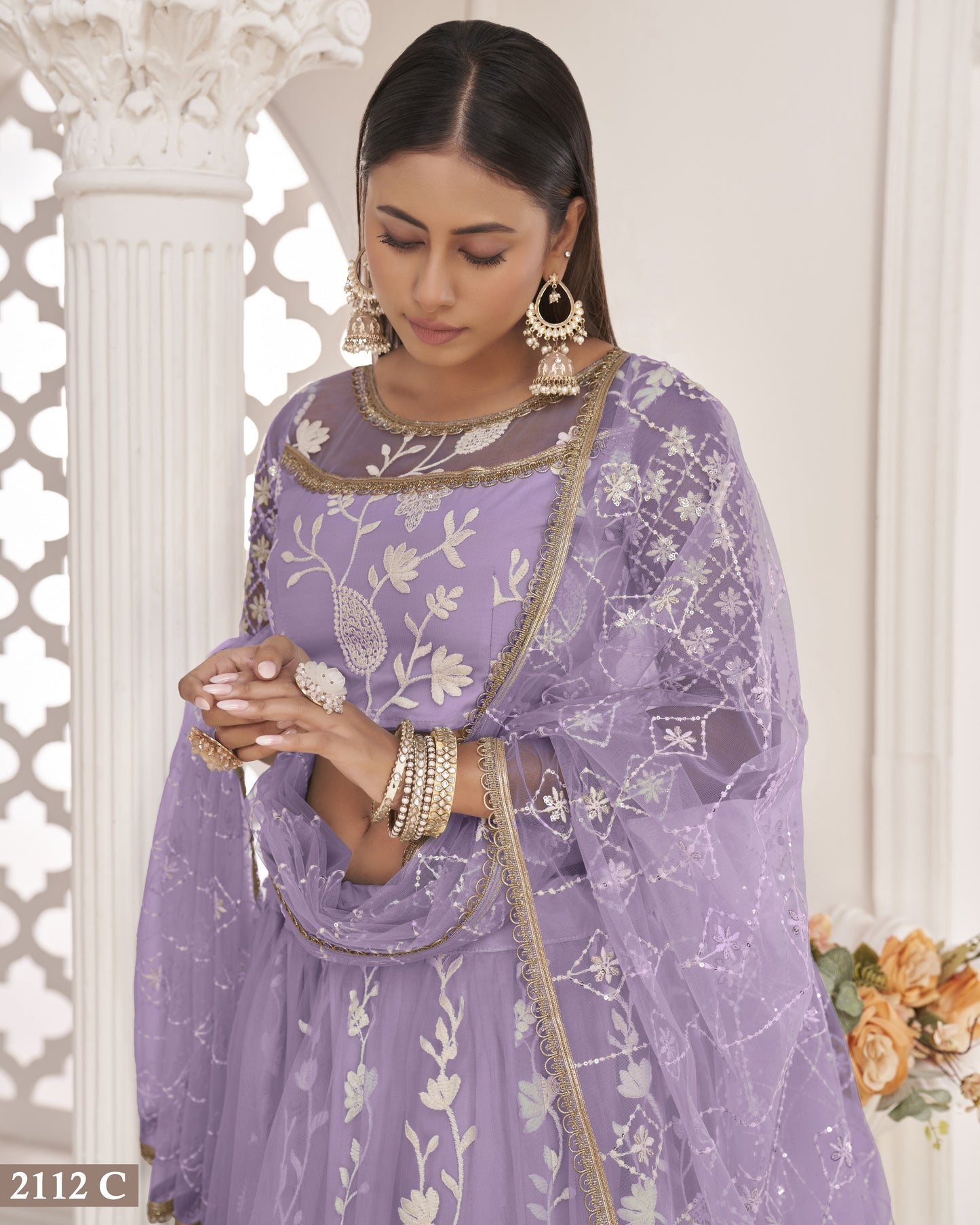 Woman in a purple embroidered traditional outfit with floral patterns, standing against a white decorative wall.