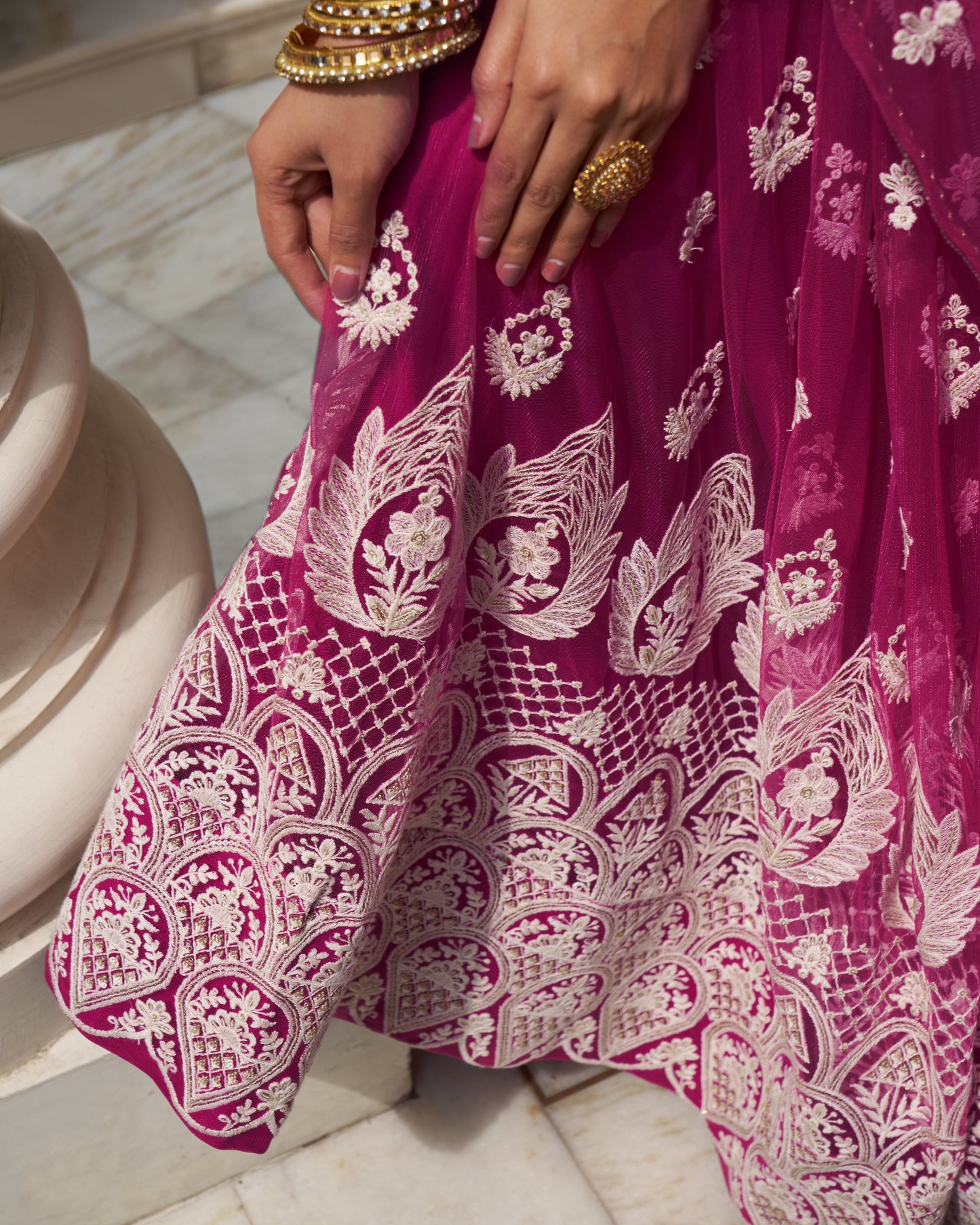 Close-up of a person wearing a vibrant pink traditional outfit with intricate white patterns, sitting on a marble floor.