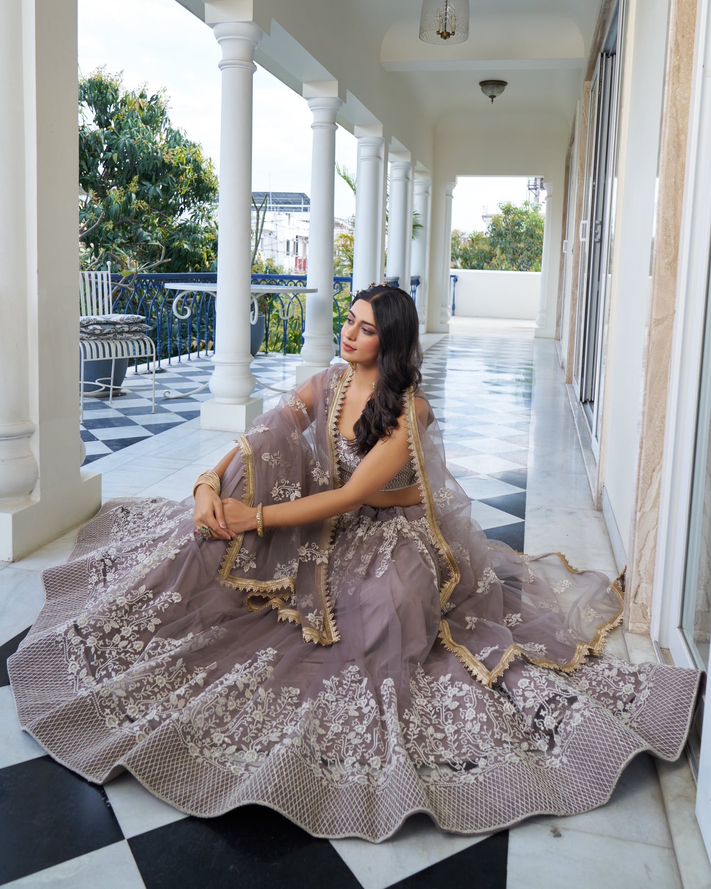 Woman in a traditional outfit sitting on a checkered floor with columns and greenery in the background