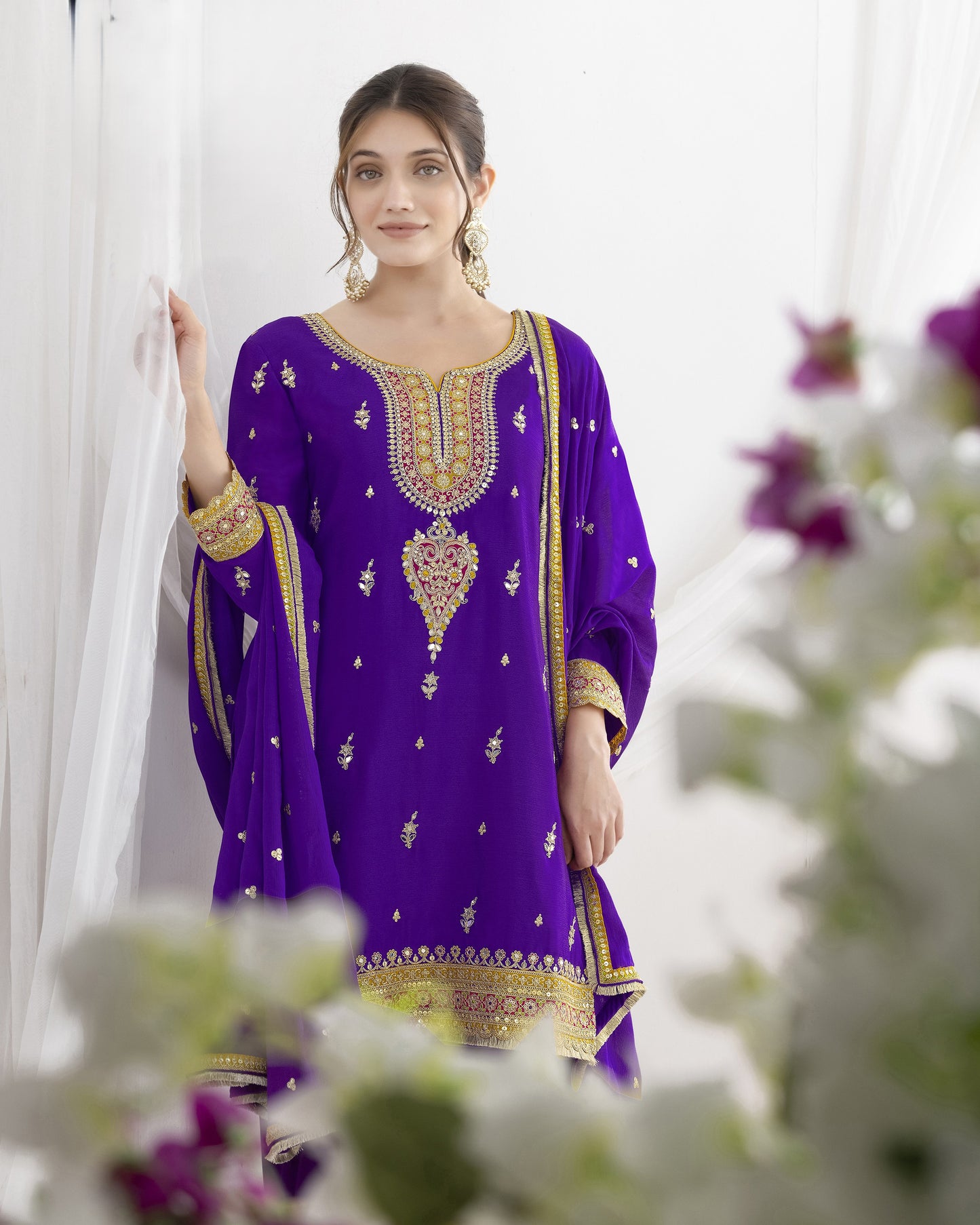 Woman wearing a purple embroidered dress with gold details against a white curtain background.