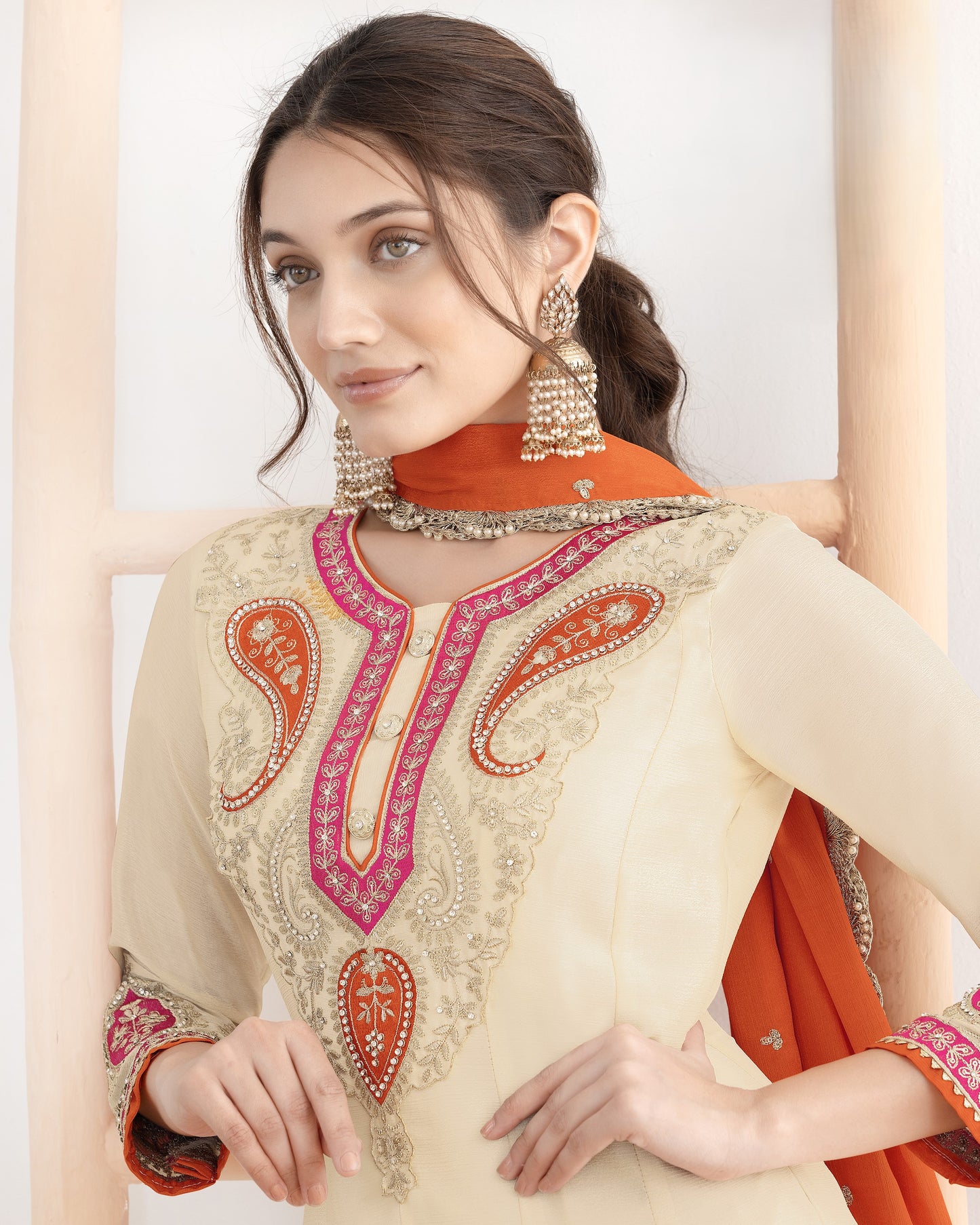 Woman wearing a cream embroidered outfit with orange dupatta against a white background