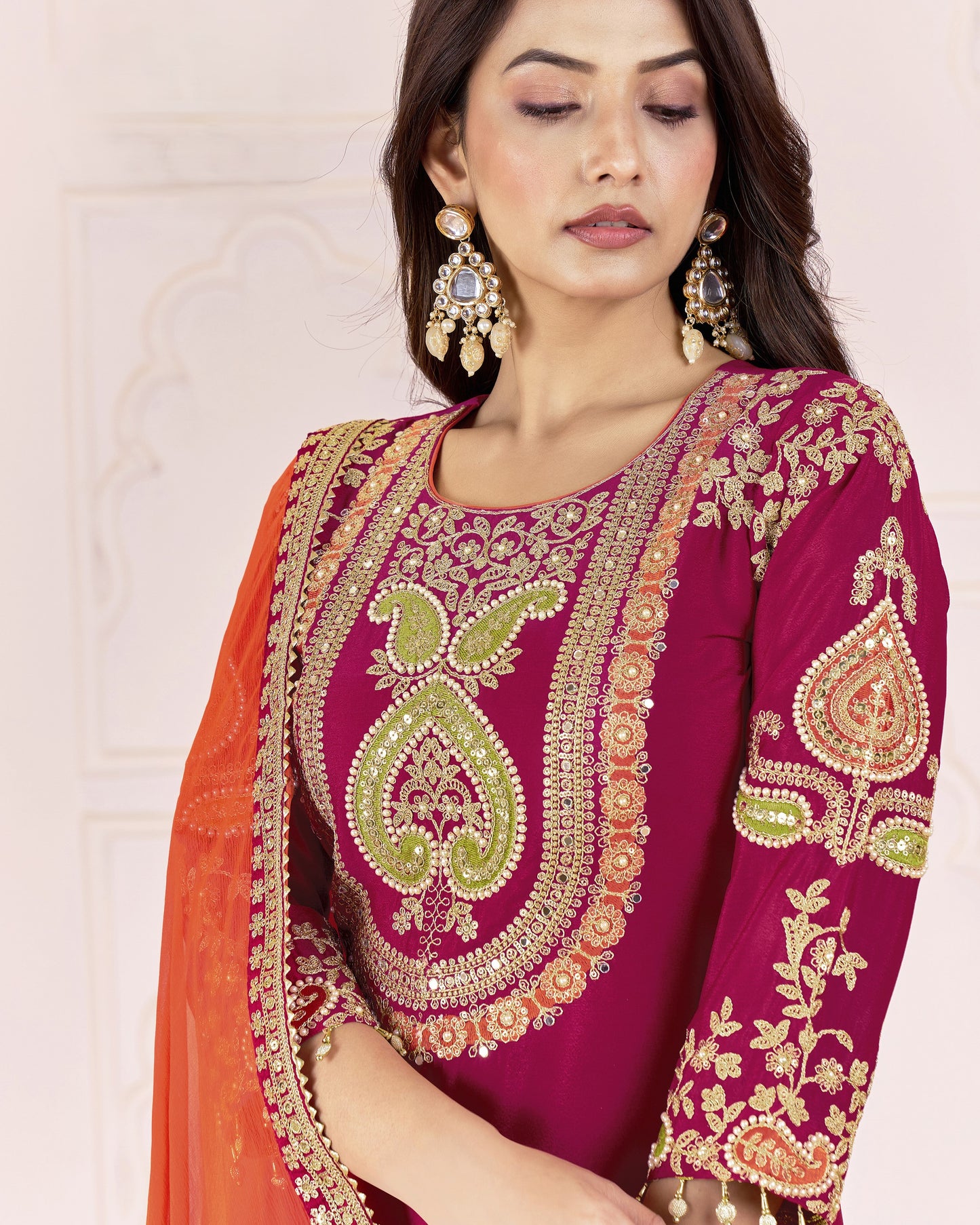 Woman wearing a maroon and orange traditional outfit with intricate embroidery on a beige background