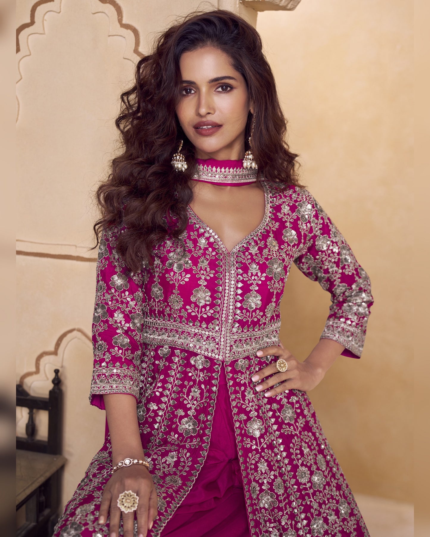 Woman wearing a pink embroidered traditional outfit with jewelry against a beige wall.