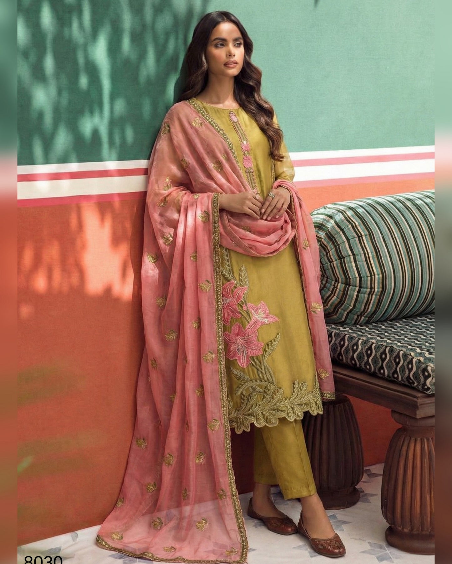 Woman in a yellow outfit with pink dupatta standing against a colorful wall.