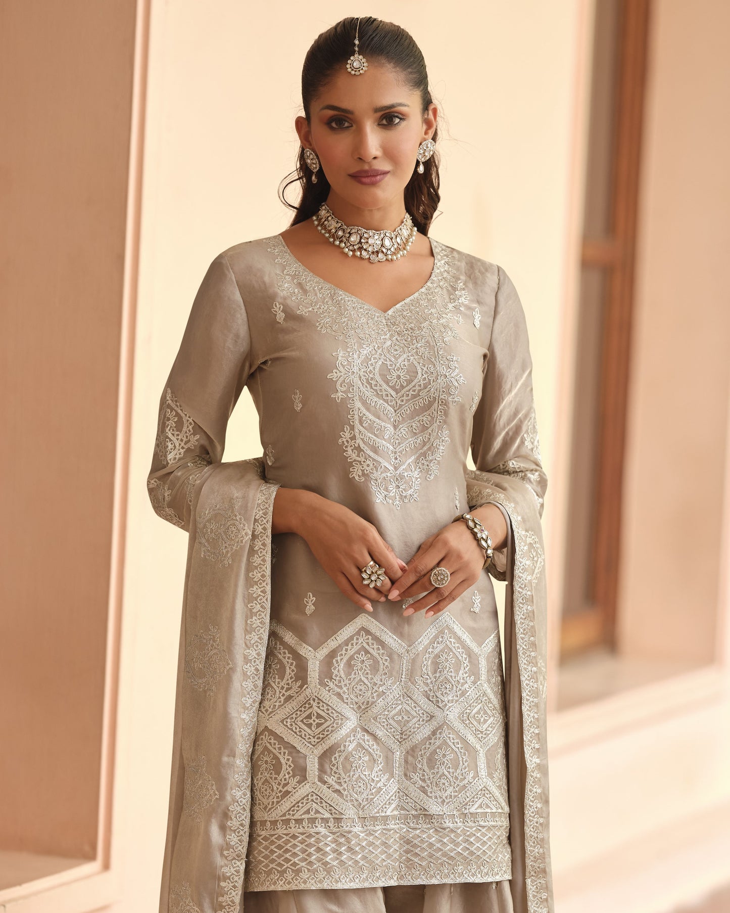 Woman wearing a light gray embroidered traditional outfit with jewelry indoors.