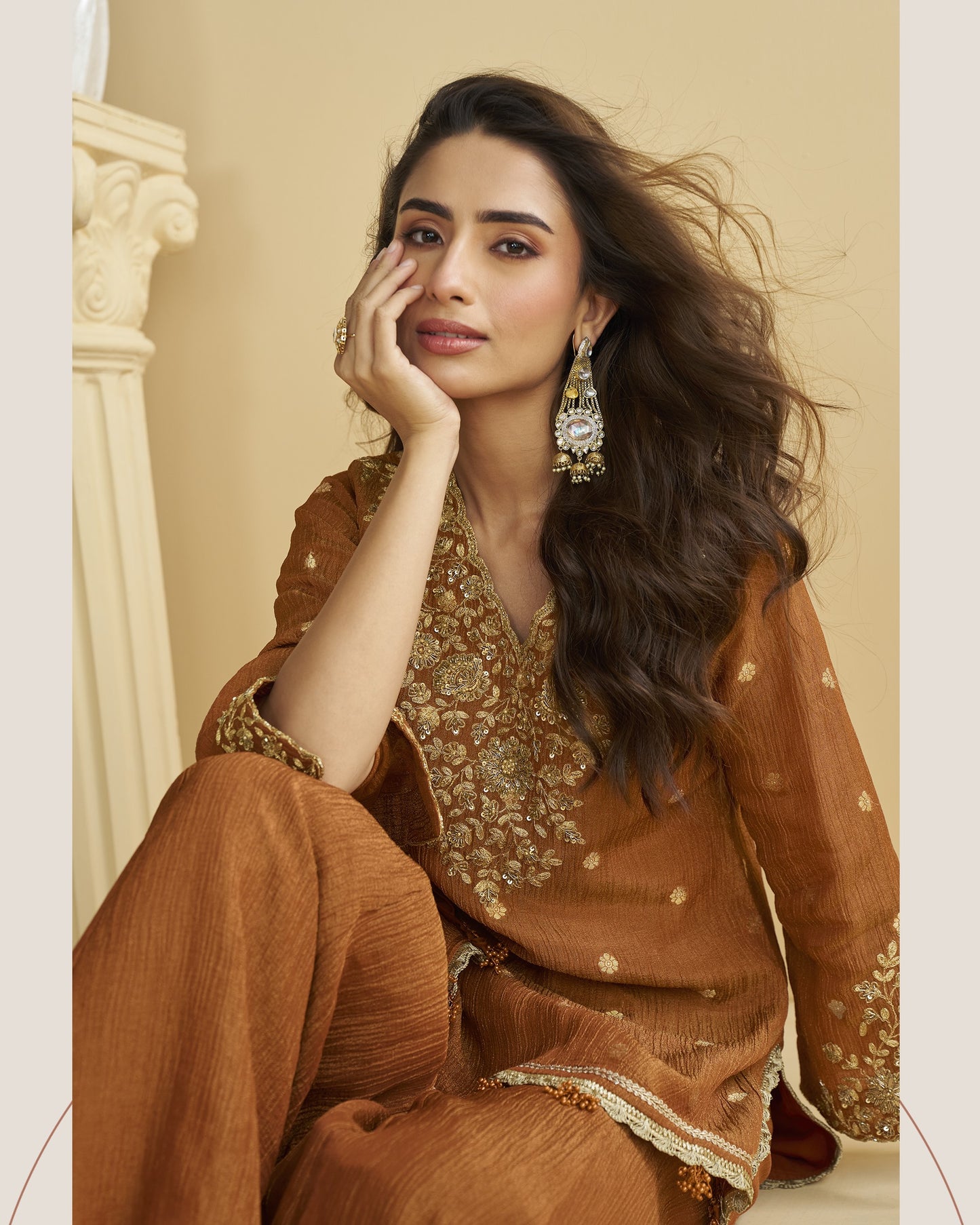 Woman in a brown traditional outfit with gold embroidery, sitting against a beige wall.