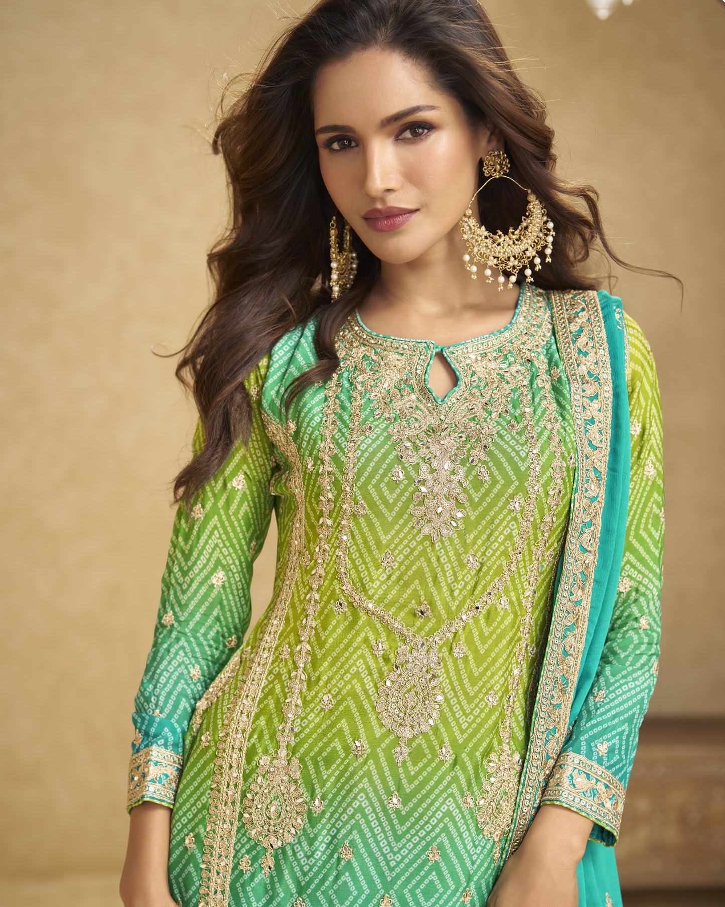 Woman wearing a green and blue embroidered traditional outfit with gold jewelry against a beige background