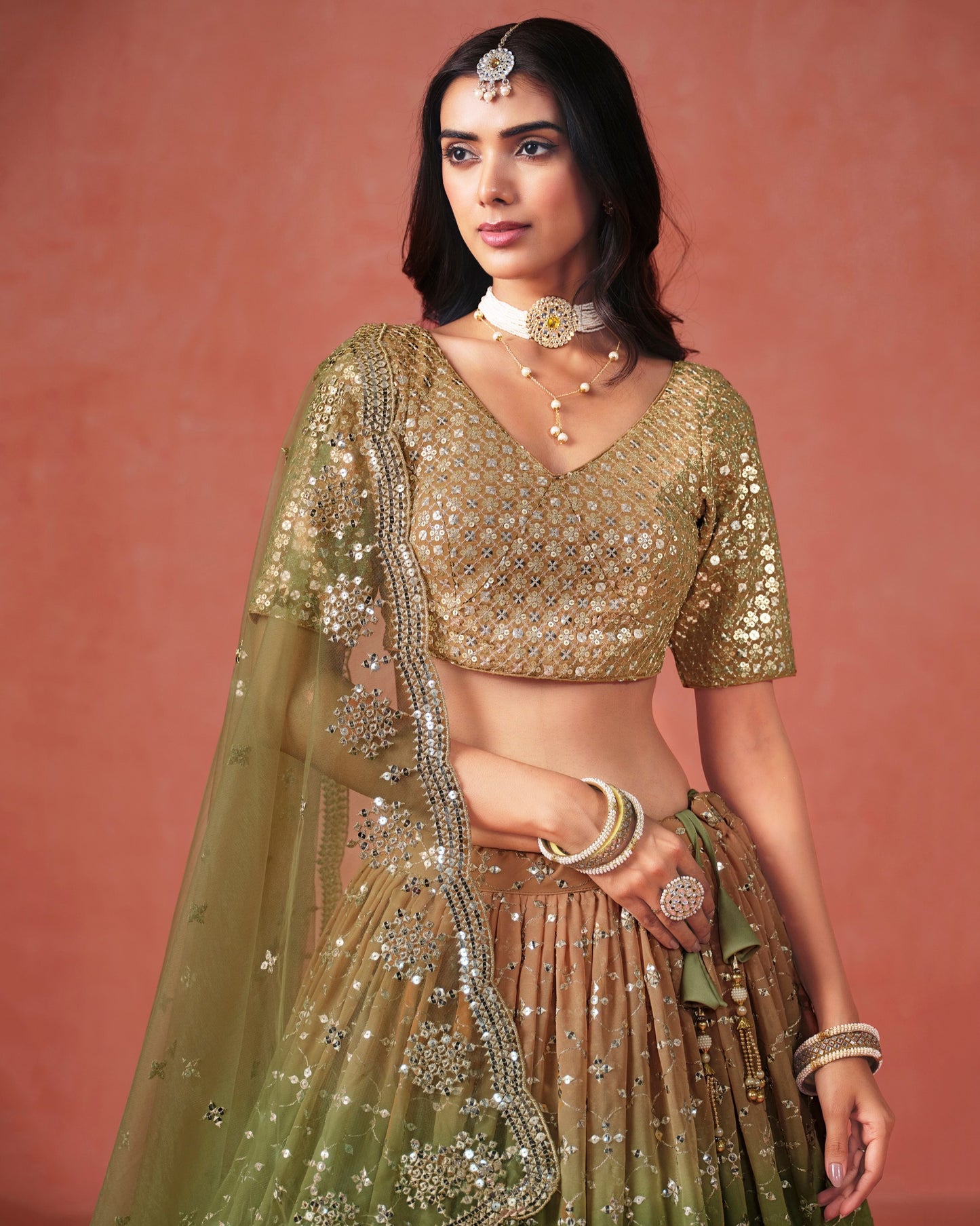 Woman wearing a traditional green and gold outfit with jewelry against a pink background