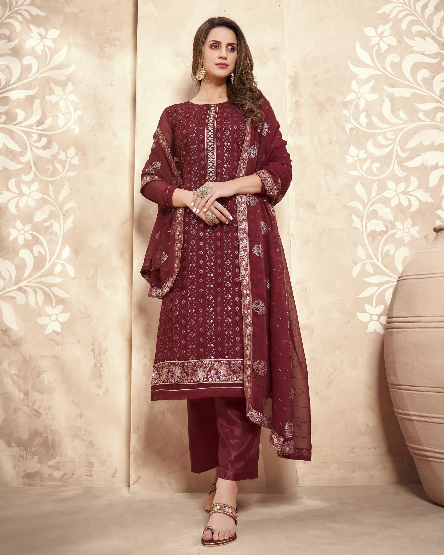Woman wearing a maroon traditional outfit with floral patterns against a decorative wall.