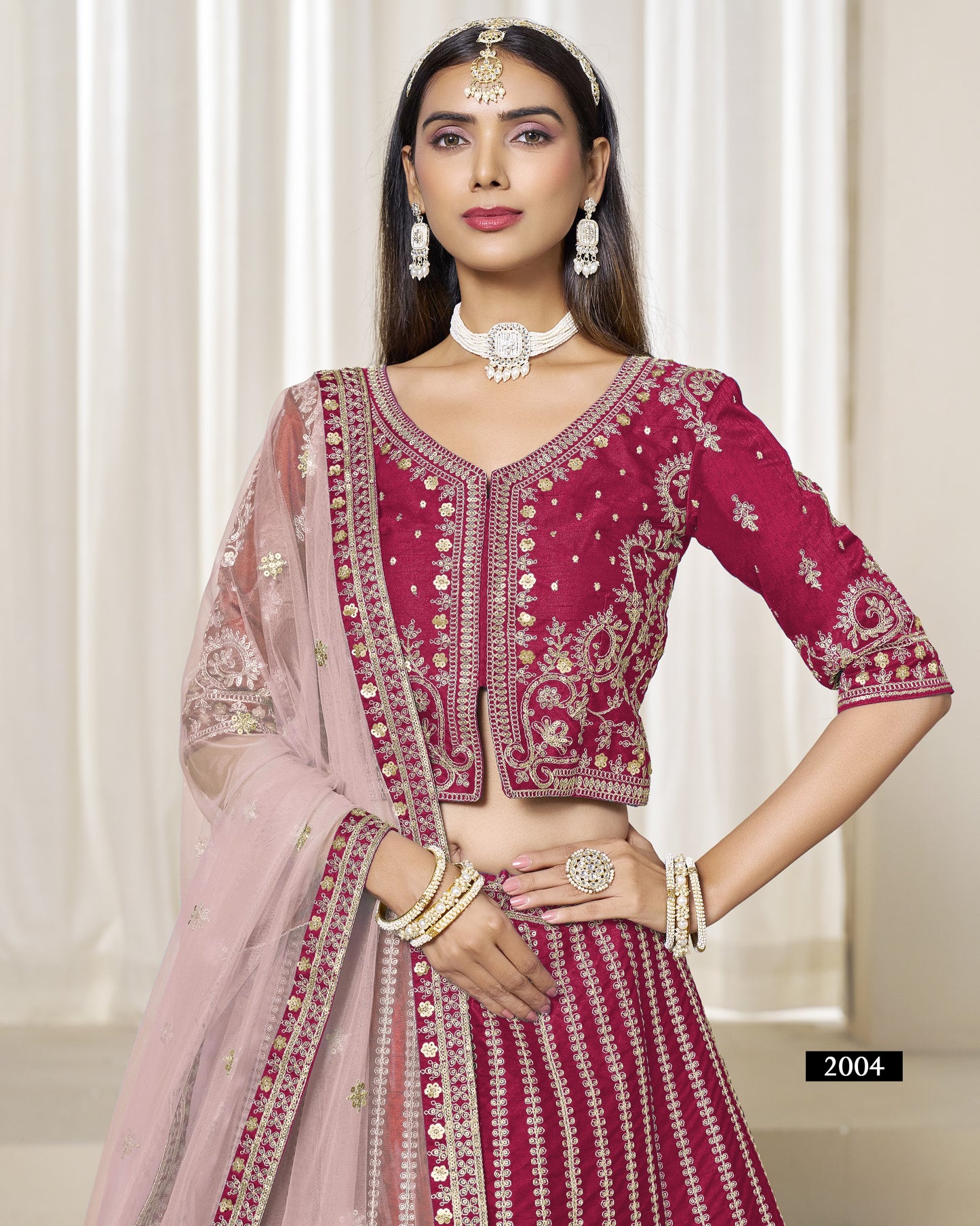 Woman wearing a traditional pink and red embroidered outfit with jewelry against a white curtain background.