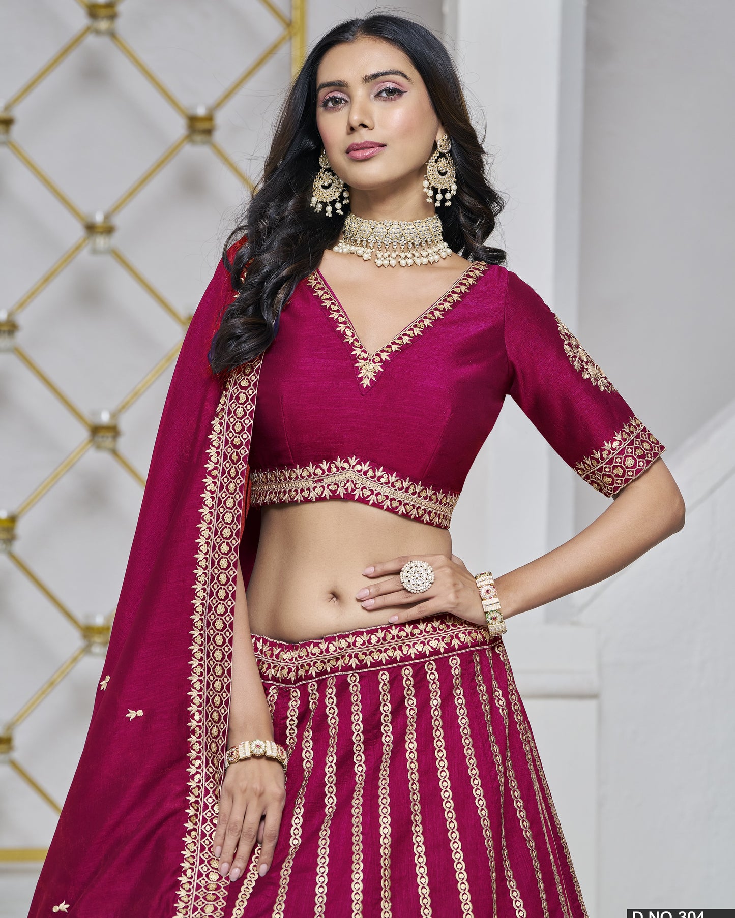 Woman wearing a maroon traditional outfit with gold embroidery against a decorative wall.