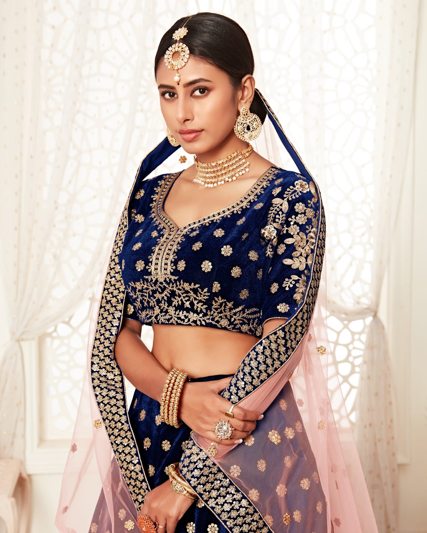 Woman in a traditional blue and gold saree with jewelry against a white curtain background