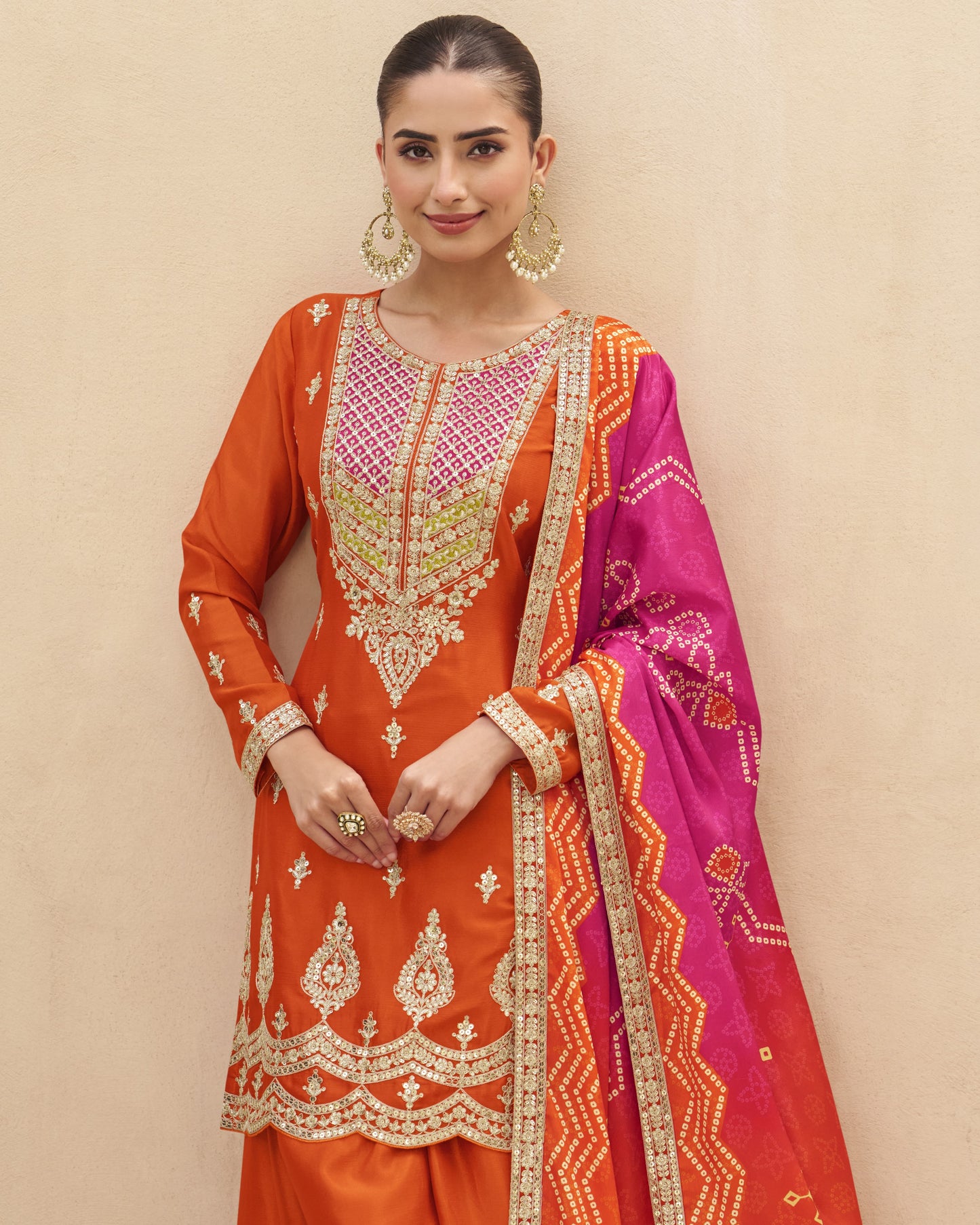 Woman wearing an orange and pink embroidered traditional outfit against a beige background