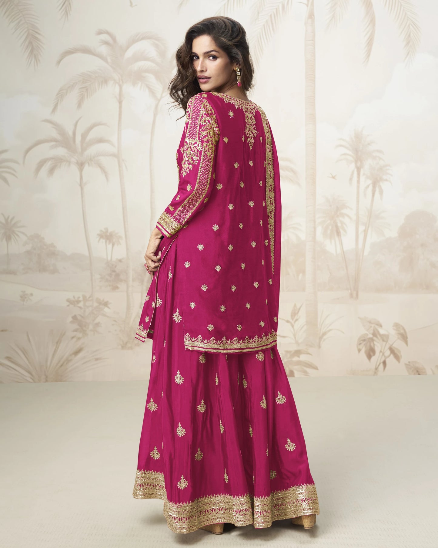 Woman wearing a pink traditional outfit with gold embroidery against a decorative wall.
