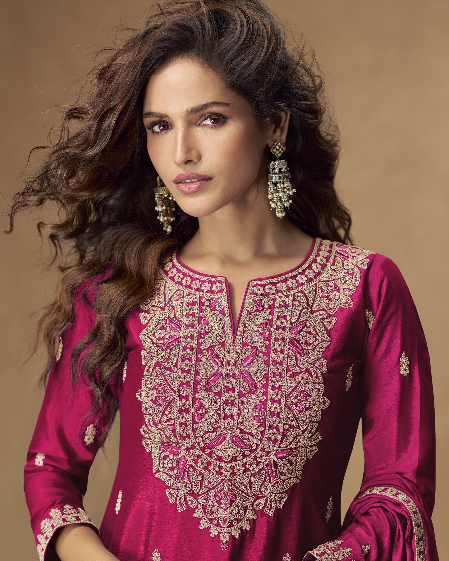Woman wearing a pink embroidered traditional outfit with gold jewelry against a brown background