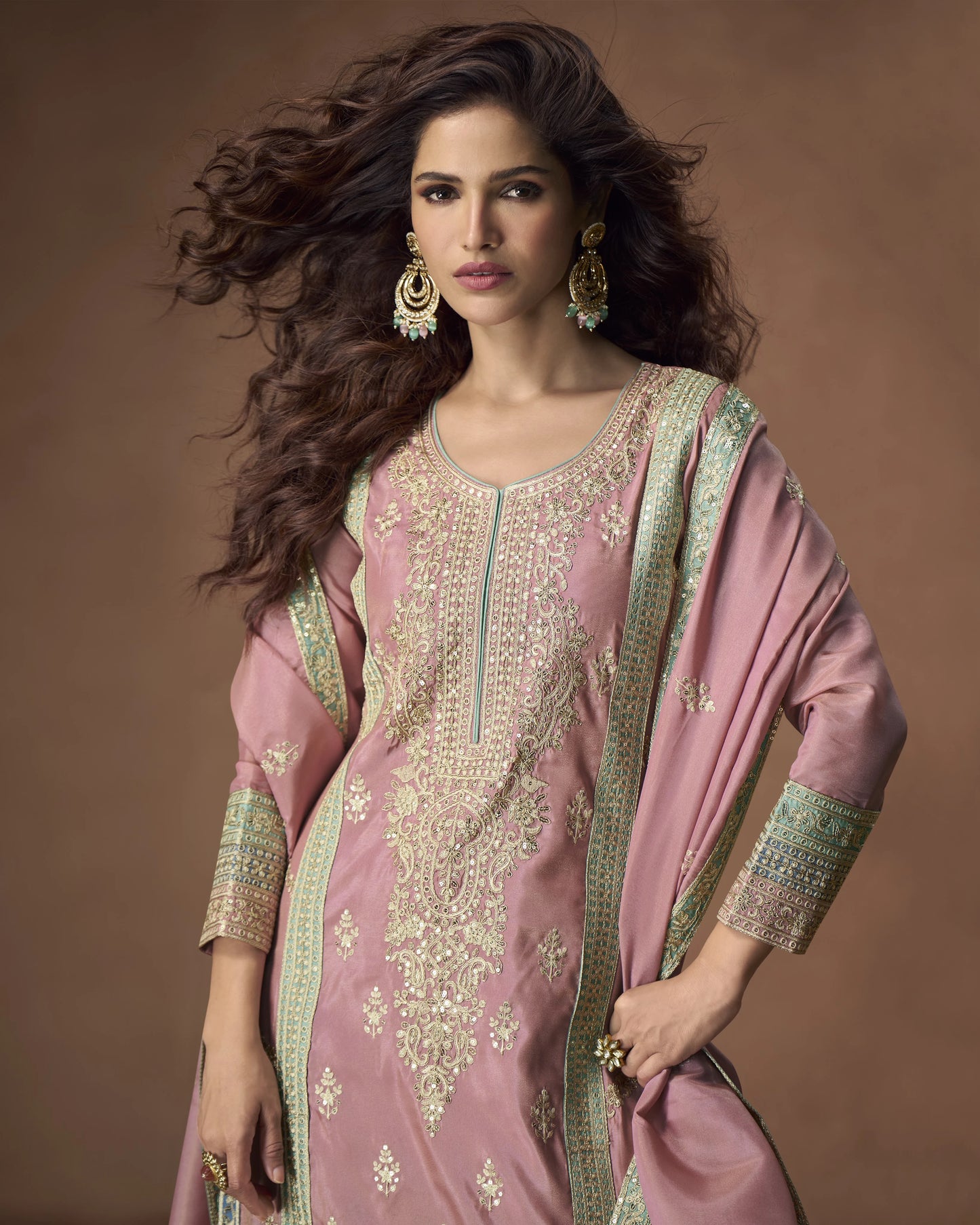 Woman wearing a pink embroidered traditional outfit with gold jewelry against a brown background