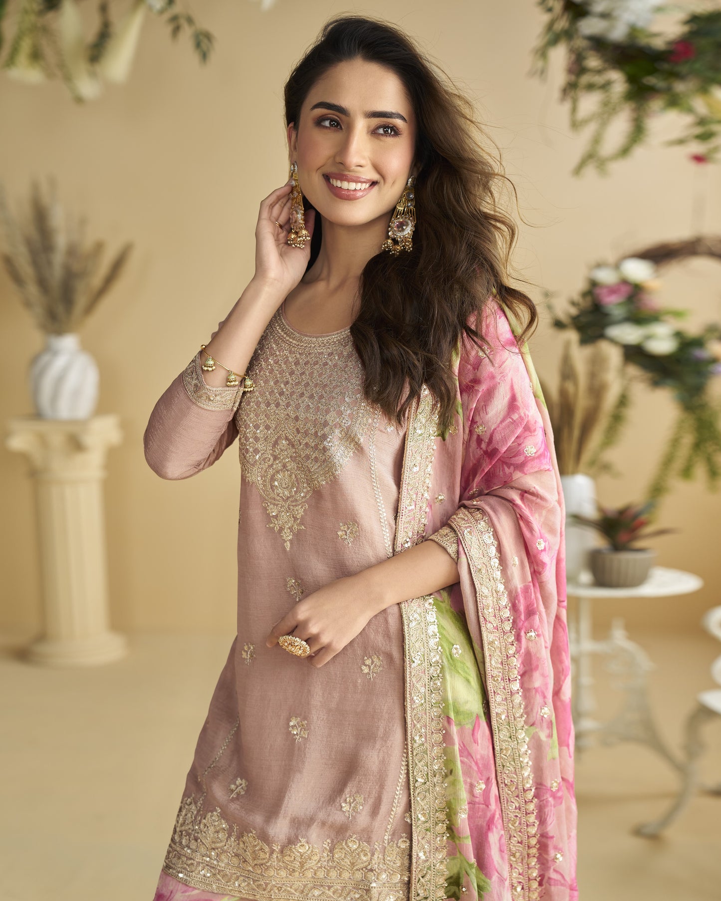Woman wearing a traditional outfit with a pink and gold dupatta in a decorated indoor setting.