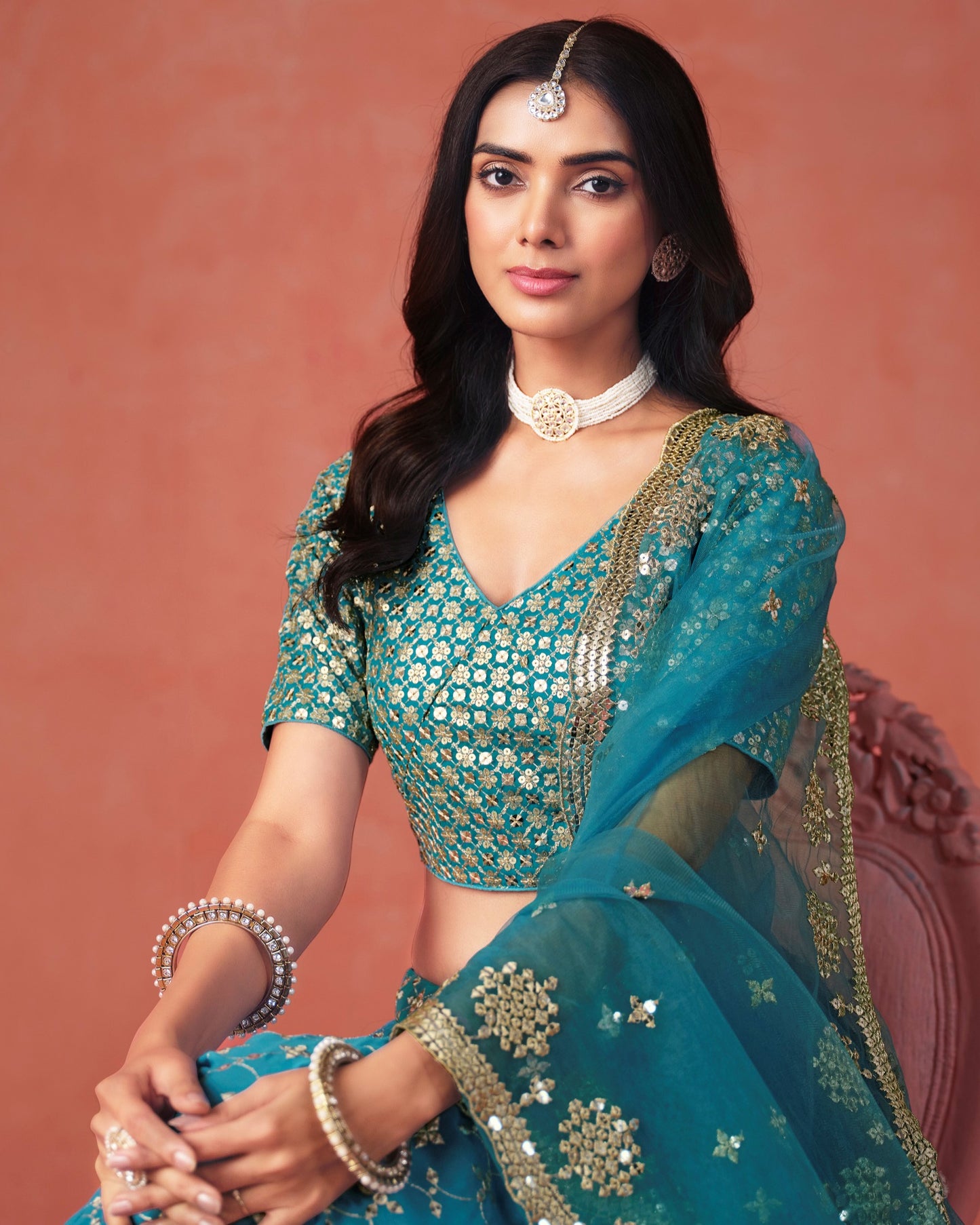 Woman wearing a traditional teal outfit with gold embroidery against a pink background