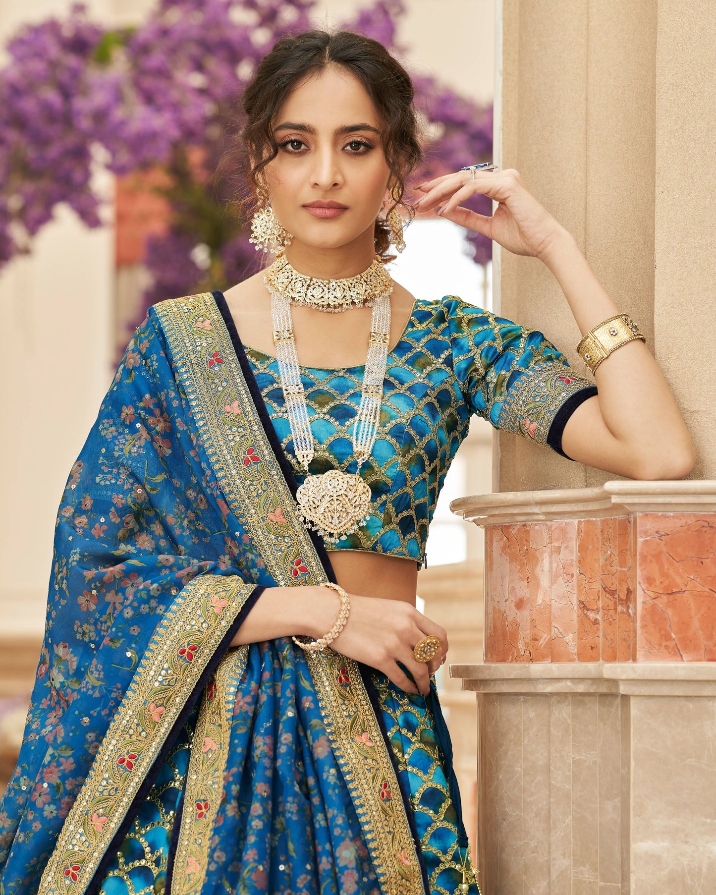 Woman in a blue saree with gold jewelry against a blurred floral background
