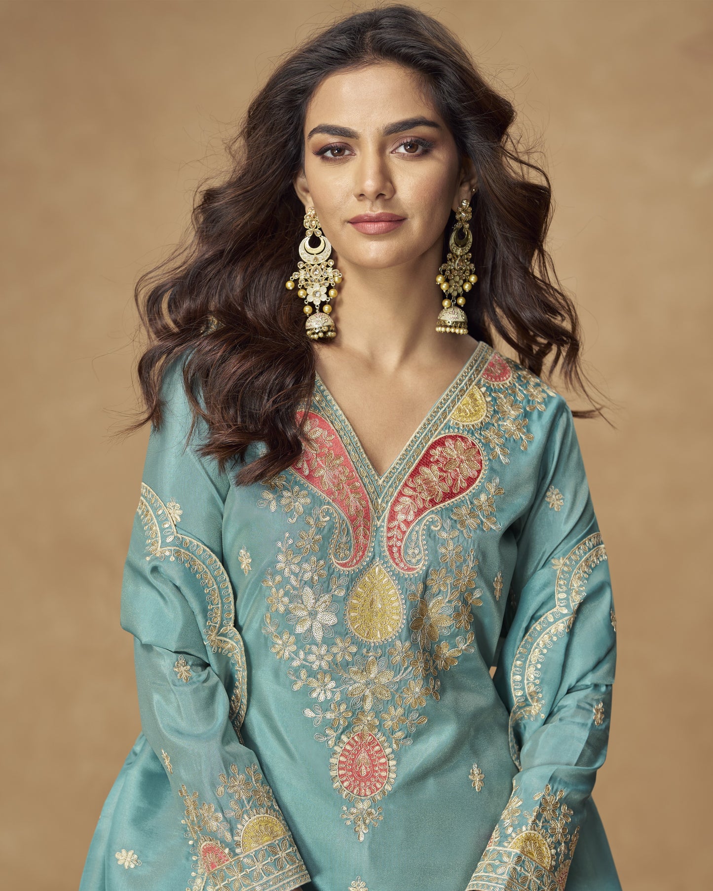 Woman wearing a teal embroidered traditional outfit with gold and red details on a beige background