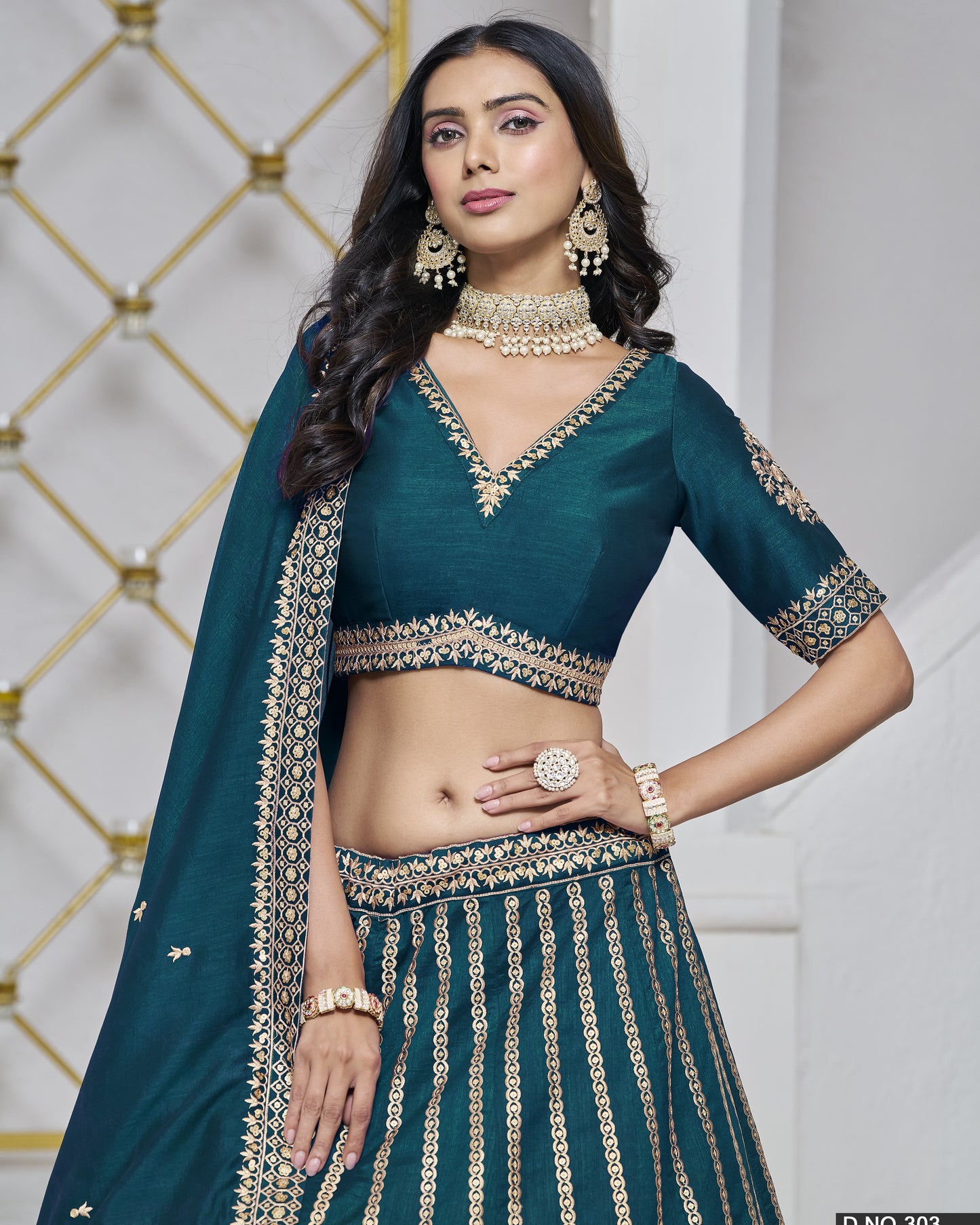 Woman wearing a teal traditional outfit with gold embroidery against a decorative wall.