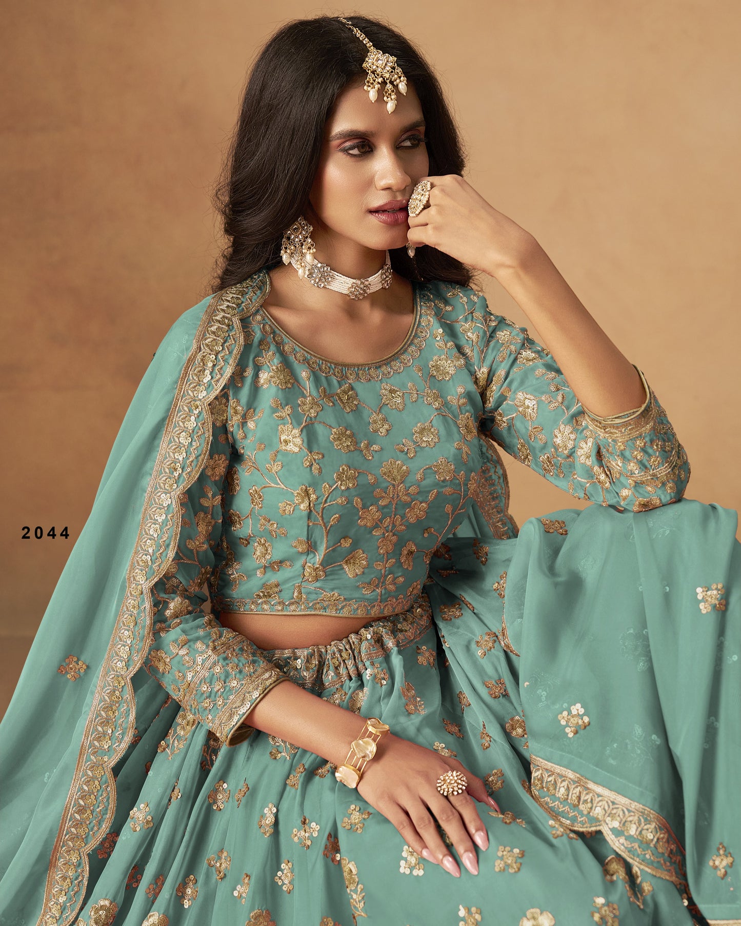 Woman in a teal traditional outfit with gold embroidery against a beige background