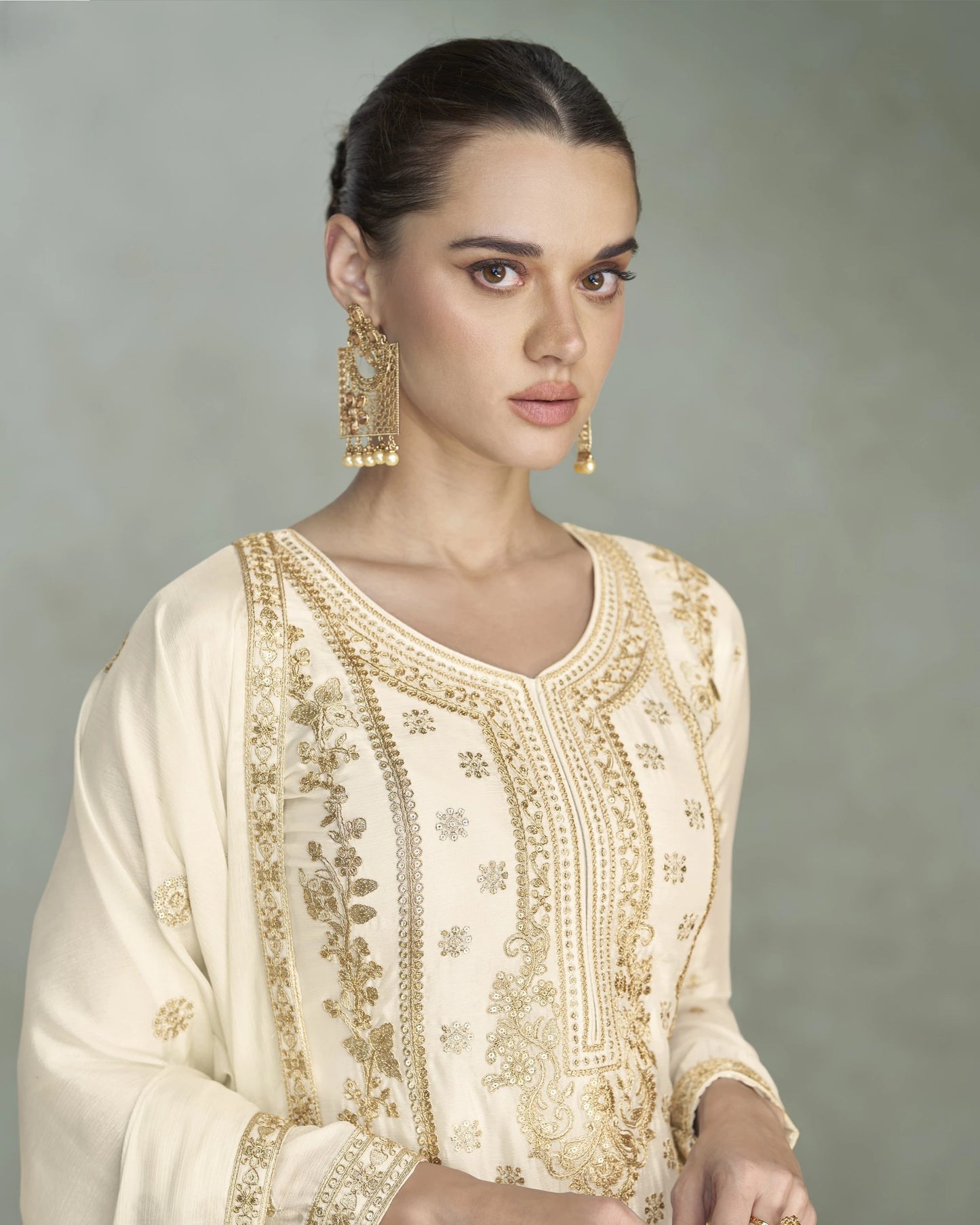 Woman wearing a cream embroidered outfit with gold details against a gray background
