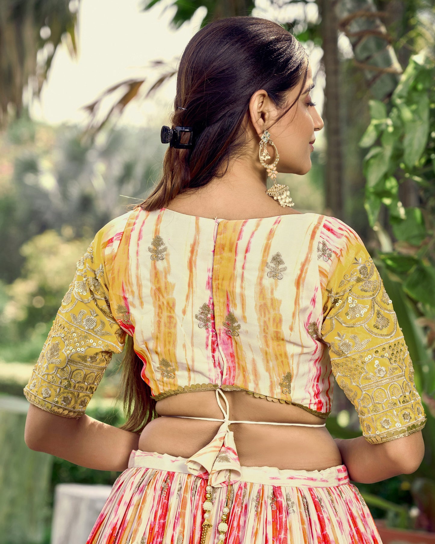 Woman wearing a traditional outfit with a colorful pattern, standing outdoors with greenery in the background.