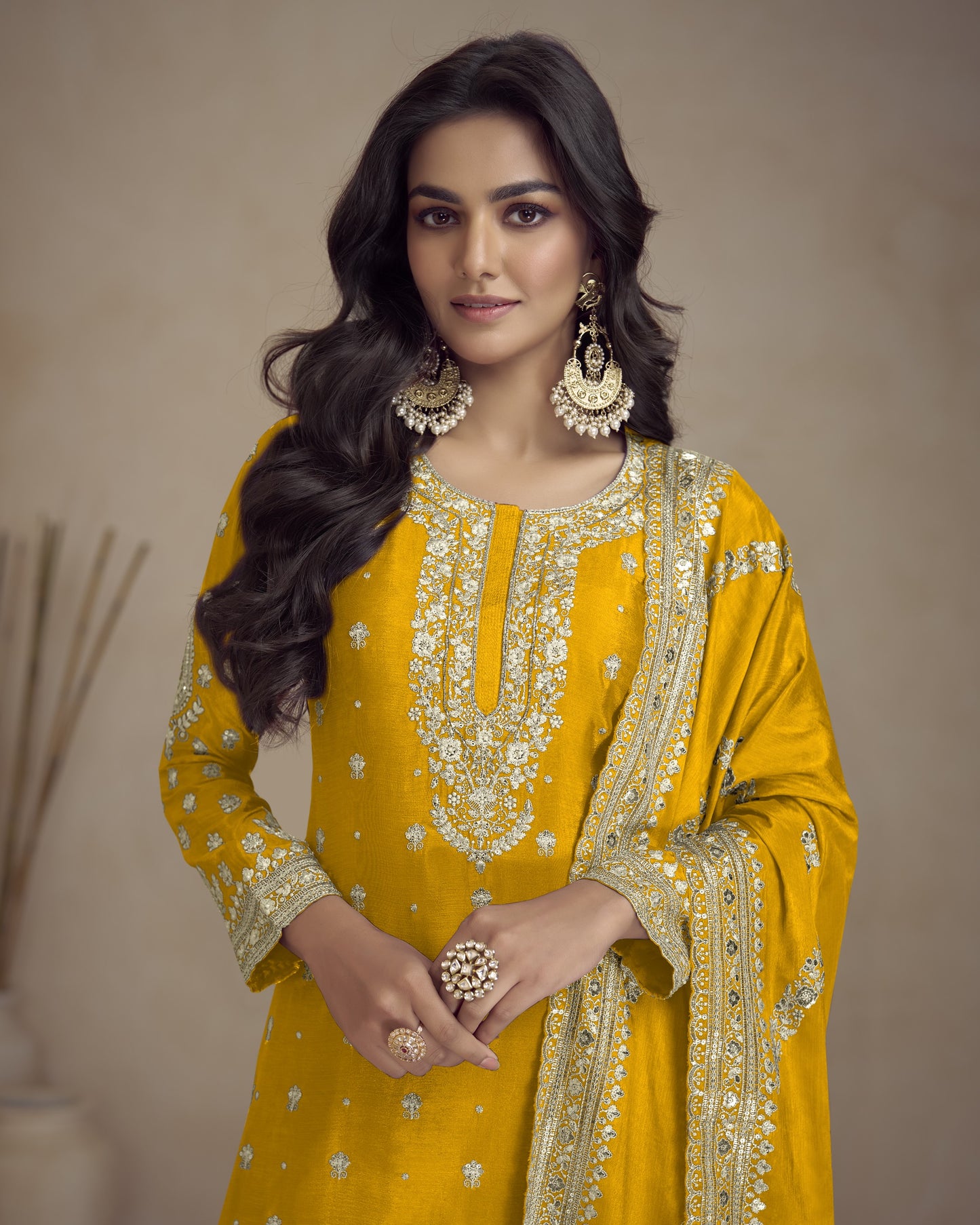 Woman wearing a yellow embroidered traditional outfit with gold jewelry against a neutral background