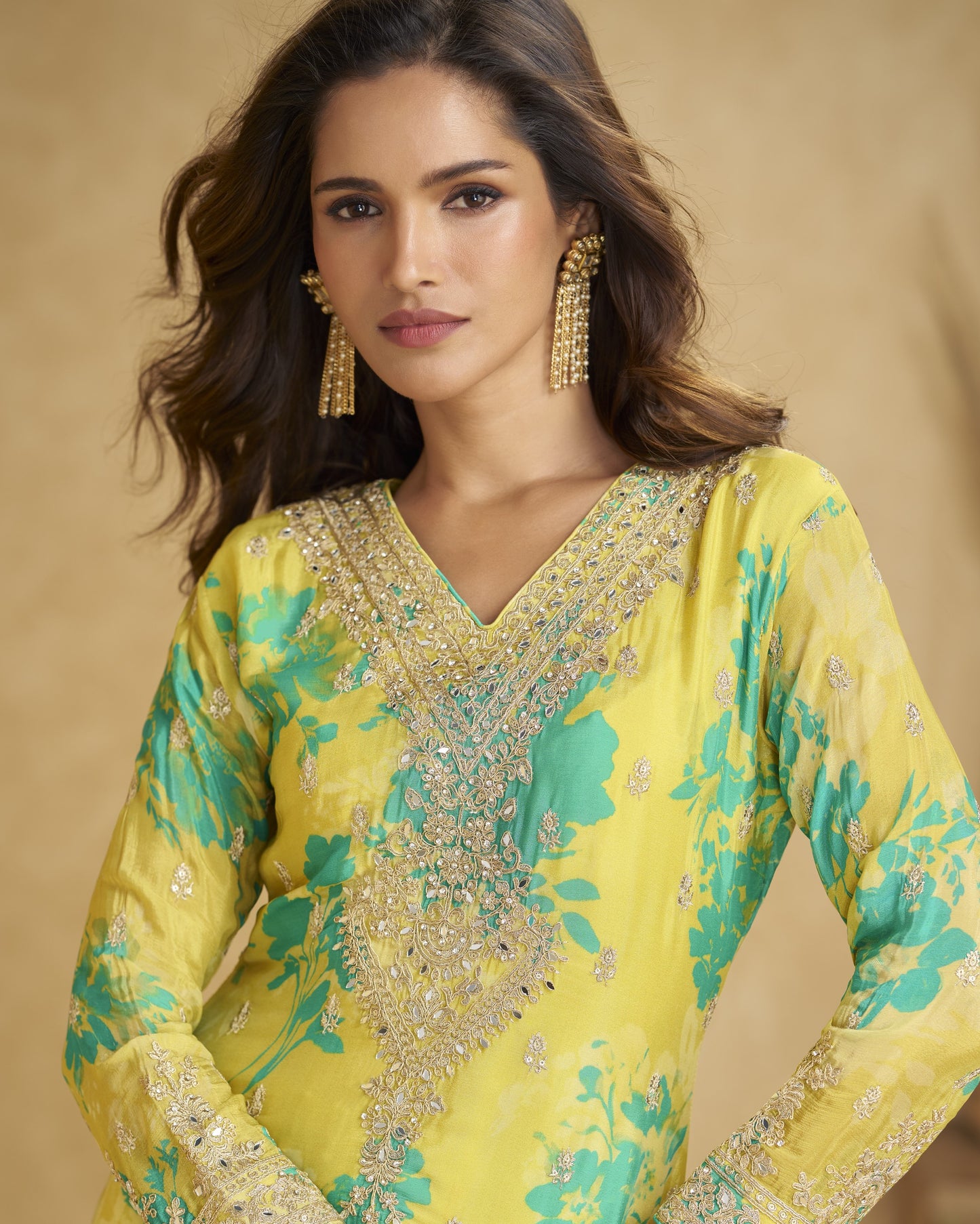 Woman wearing a yellow and green embroidered traditional outfit with gold jewelry against a beige background