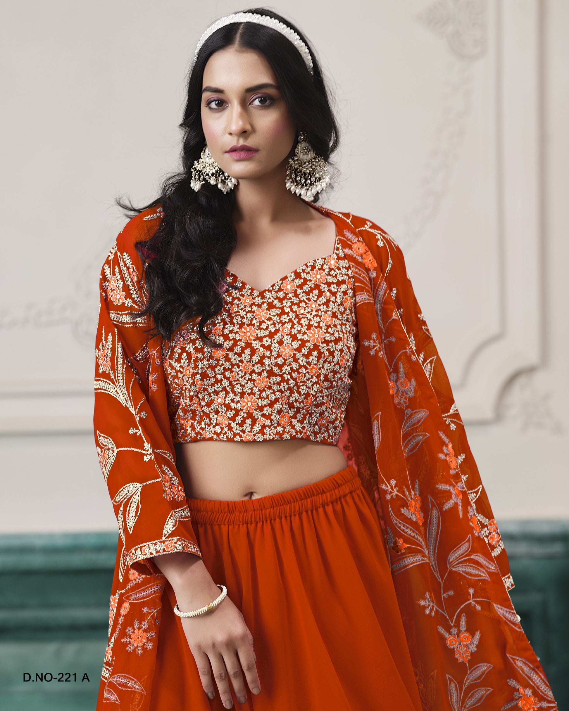 Woman wearing an orange traditional outfit with floral patterns against a decorative wall.