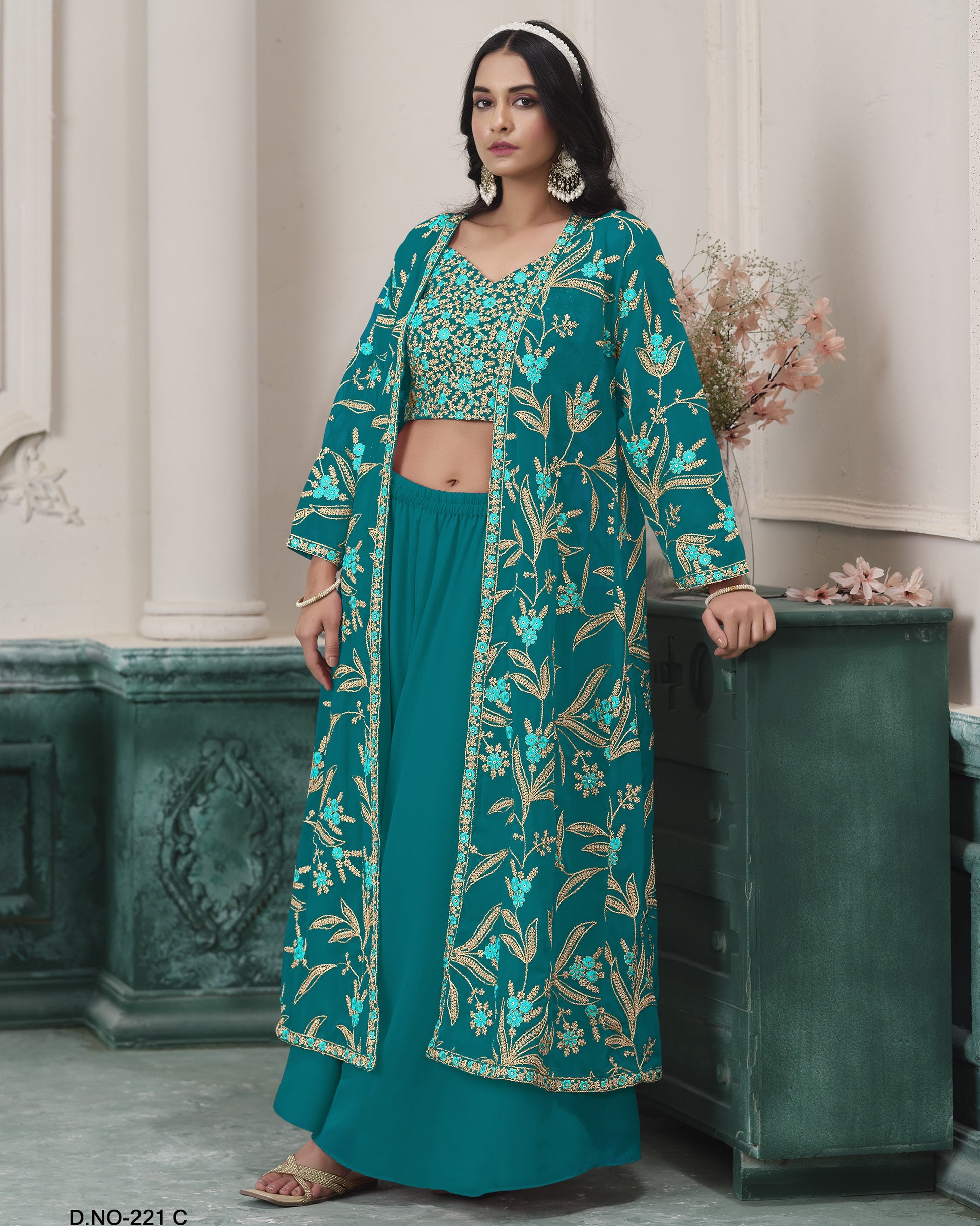 Woman wearing a teal embroidered outfit standing against a decorative wall.