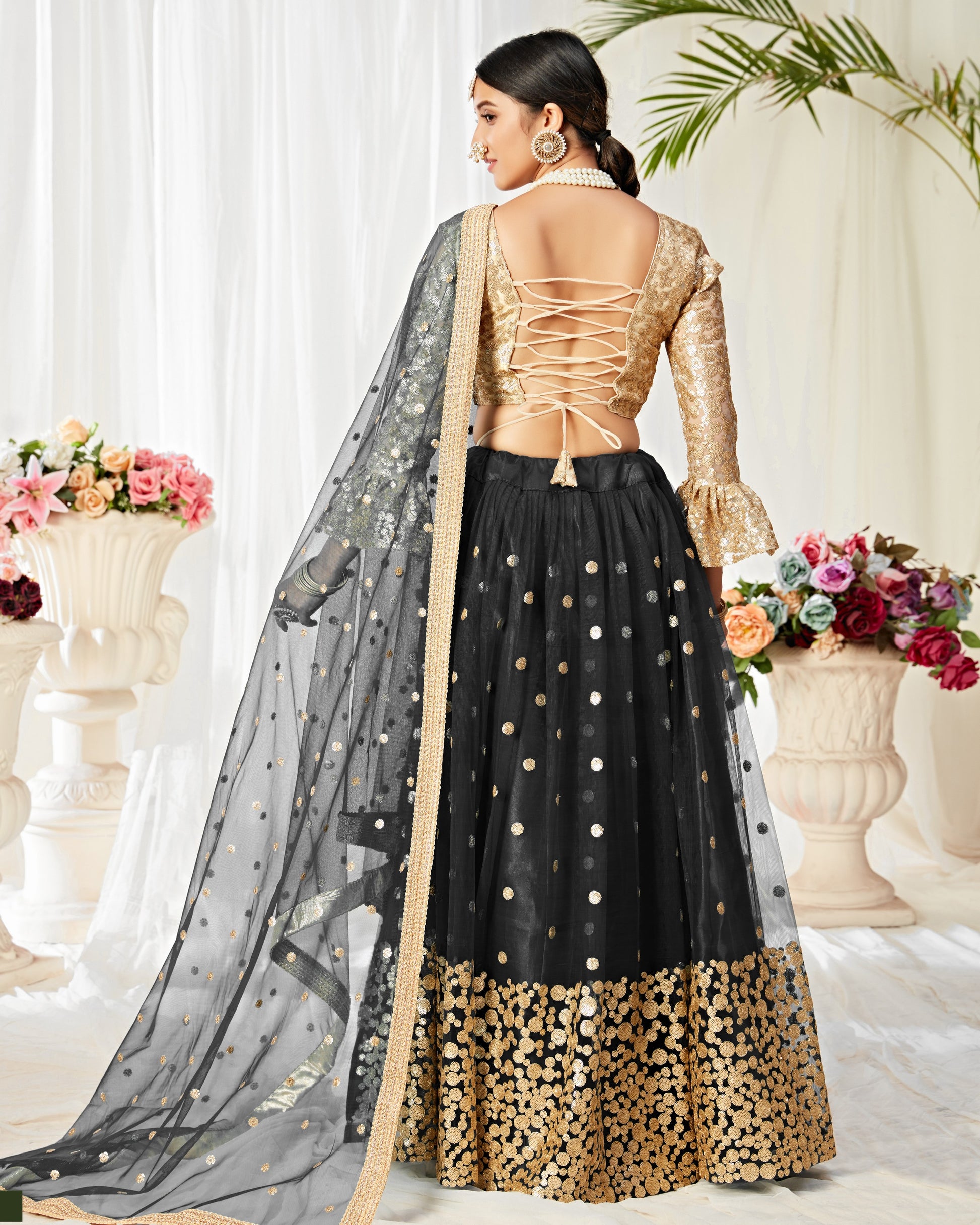 Woman wearing a black and gold traditional outfit with a sheer dupatta in a decorated indoor setting.