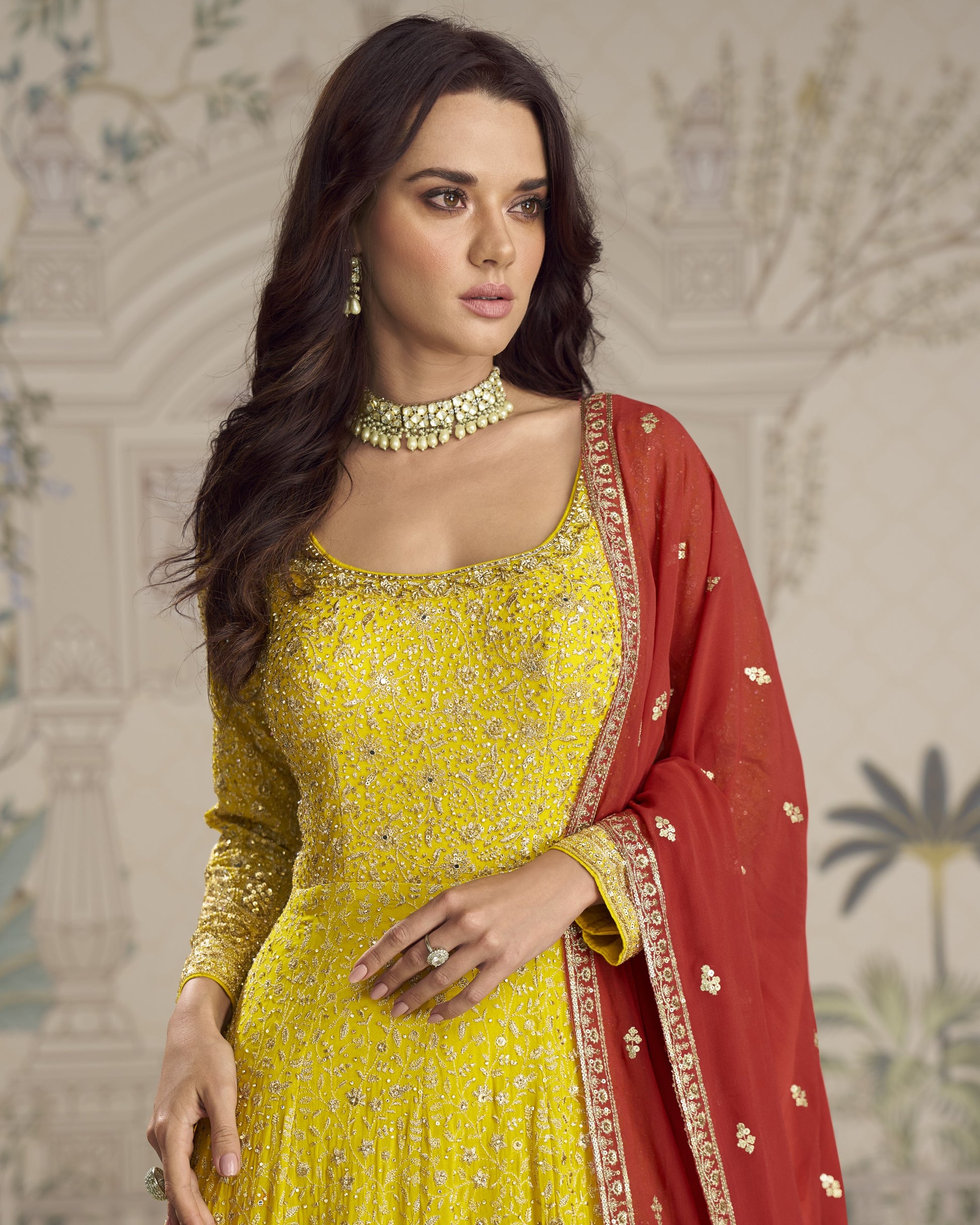 Woman in a yellow and red traditional outfit with intricate patterns against a decorative background