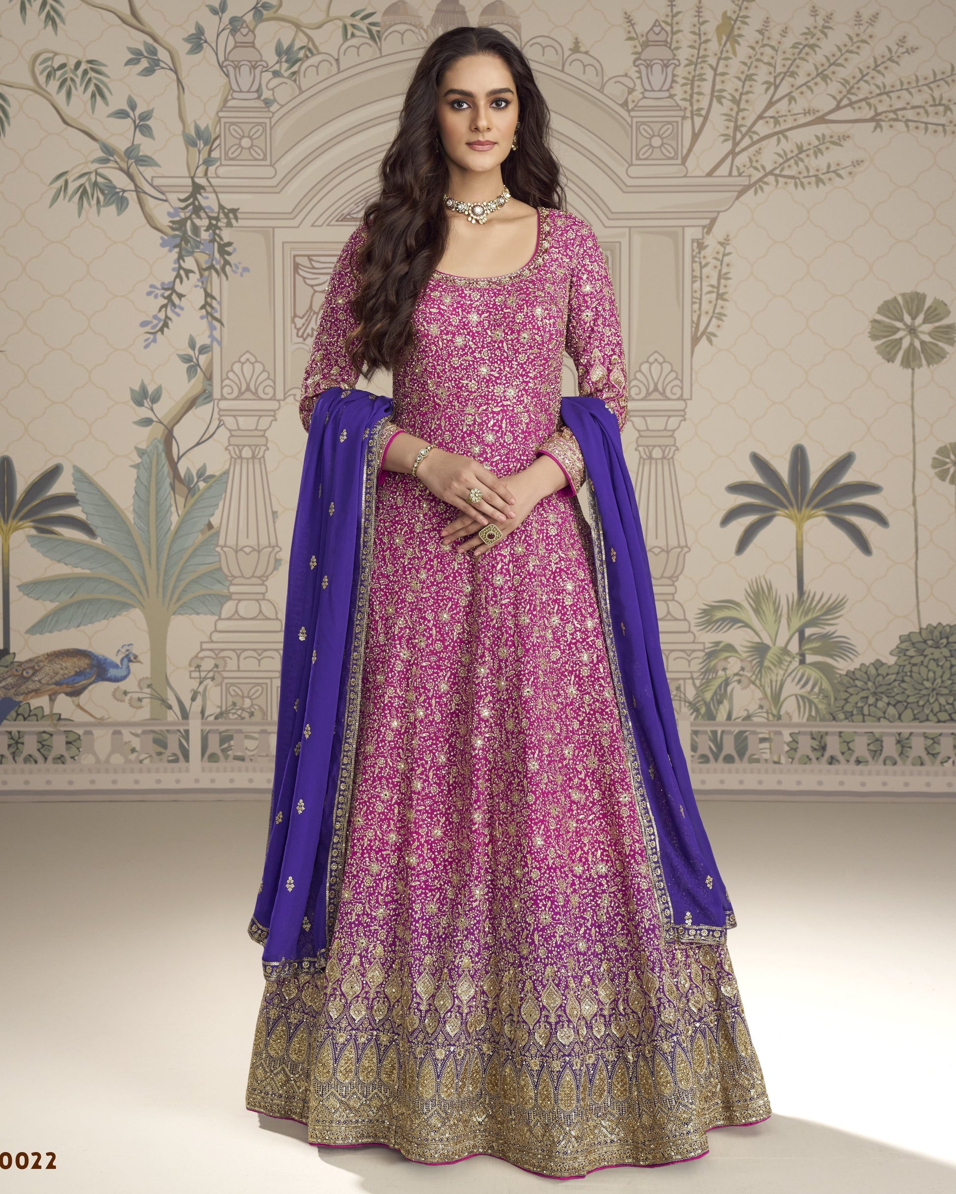 Woman wearing a pink and purple traditional outfit with a blue dupatta against a decorative wall.