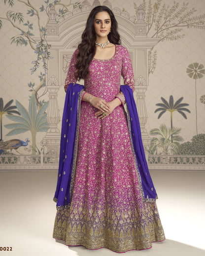 Woman wearing a pink and purple traditional outfit with a blue dupatta against a decorative wall.