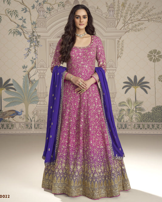 Woman wearing a pink and purple traditional outfit with a blue dupatta against a decorative wall.
