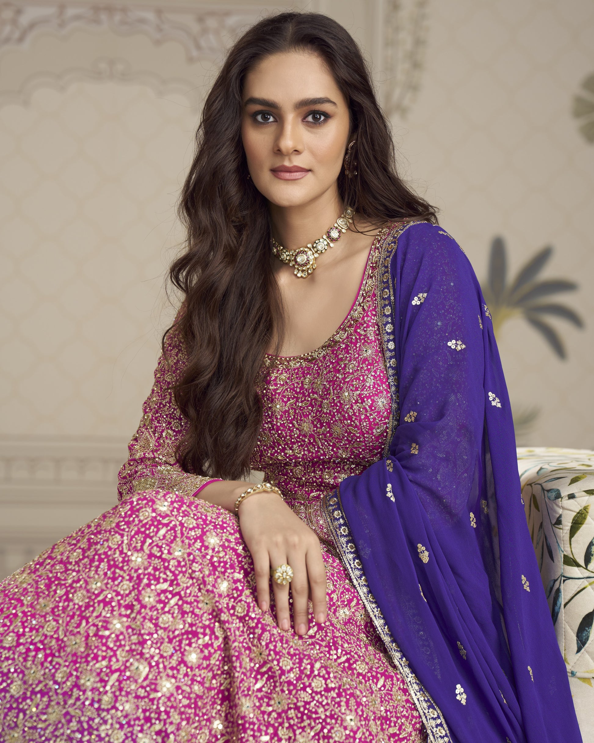 Woman in a pink and purple traditional outfit with jewelry, sitting on a decorative chair.