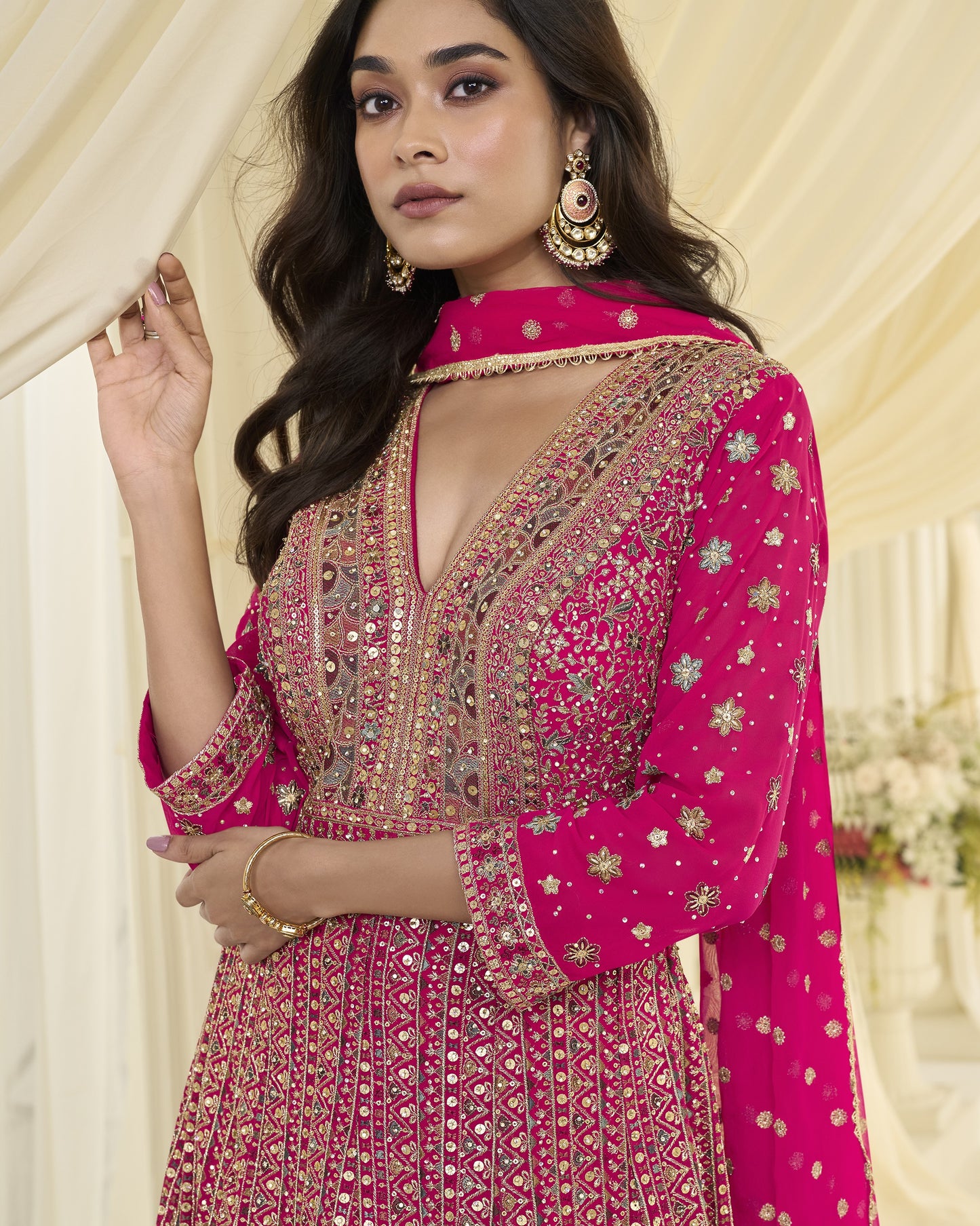 Woman wearing a pink embroidered traditional outfit with gold jewelry against a neutral background