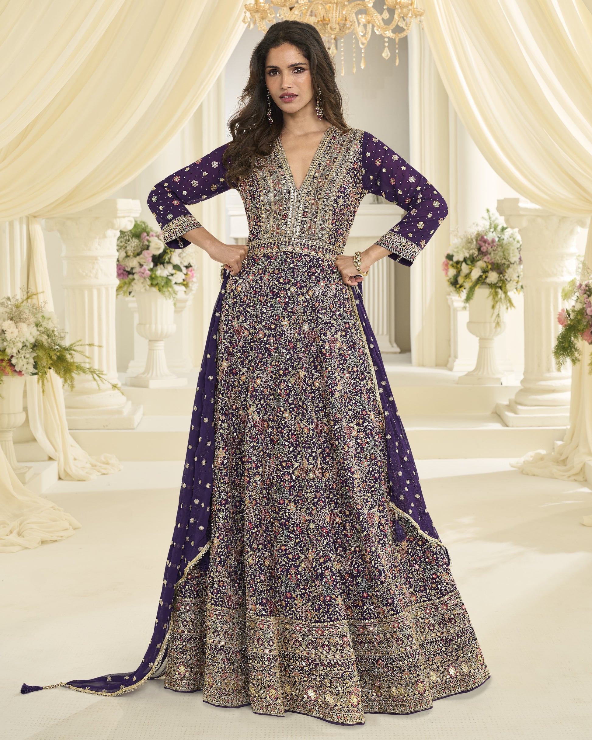 Woman in a long, patterned dress standing in an elegant indoor setting with floral arrangements and chandeliers.