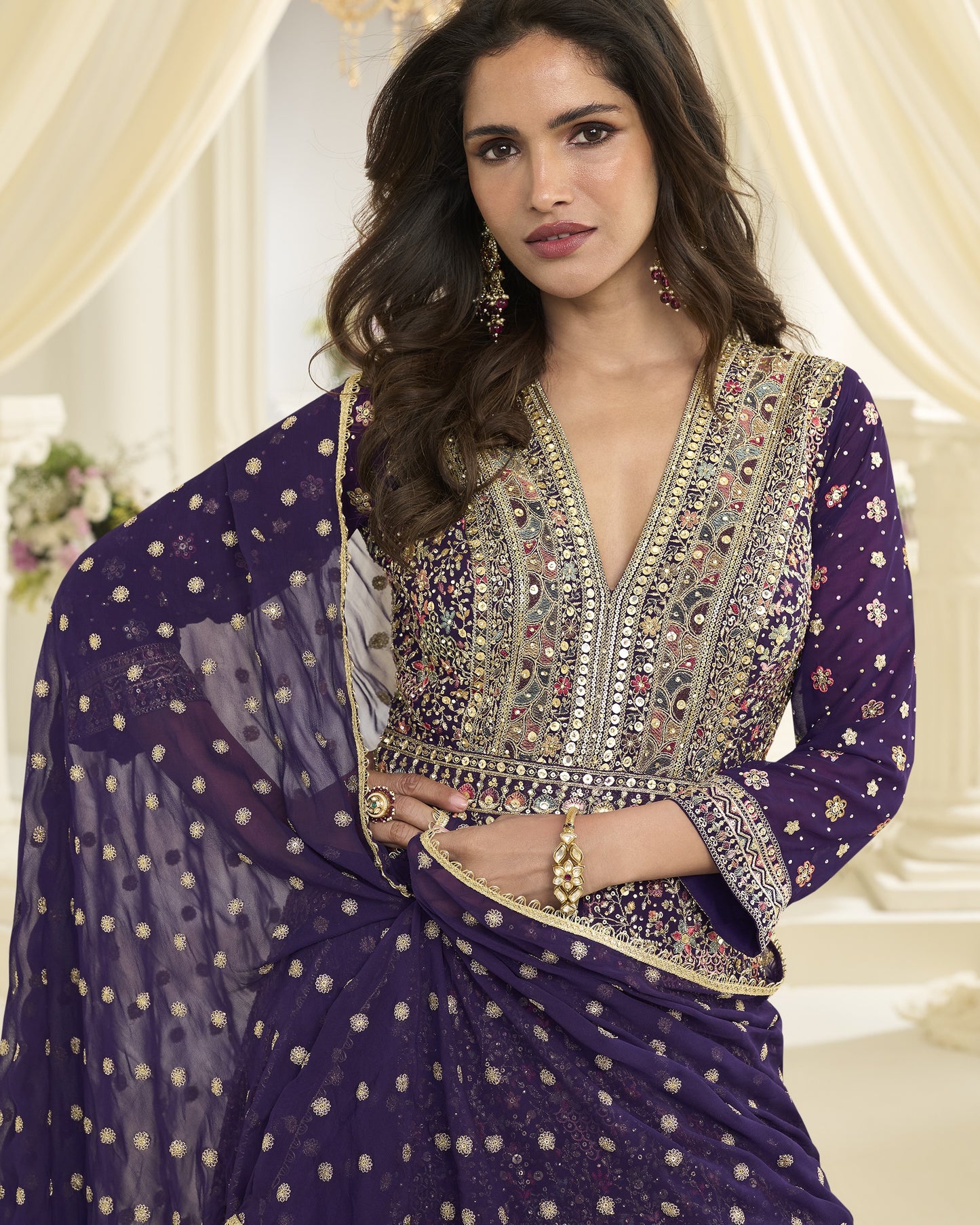 Woman wearing a traditional embroidered outfit with a purple dupatta in an elegant indoor setting.