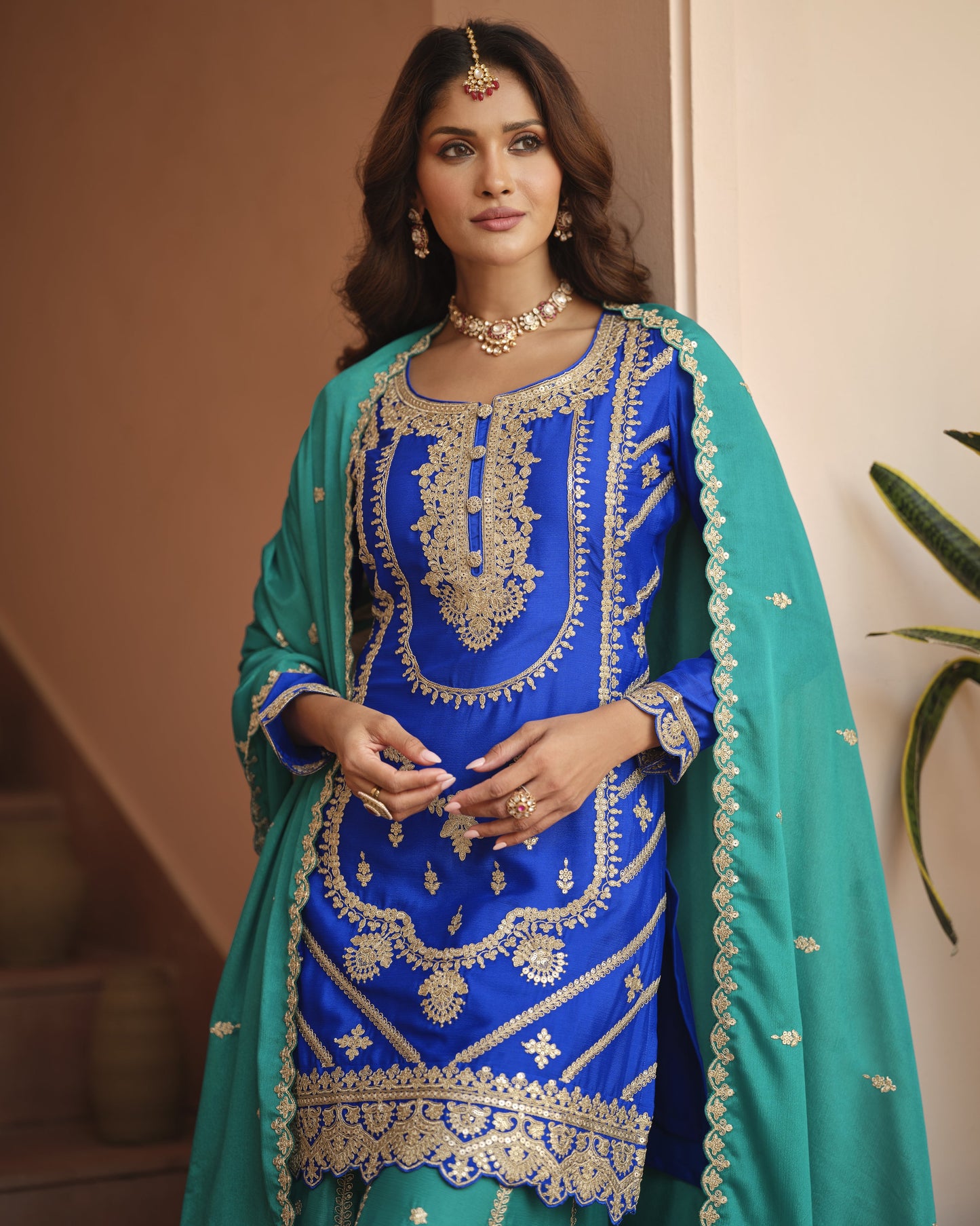 Woman wearing a blue and green embroidered traditional outfit with gold jewelry.