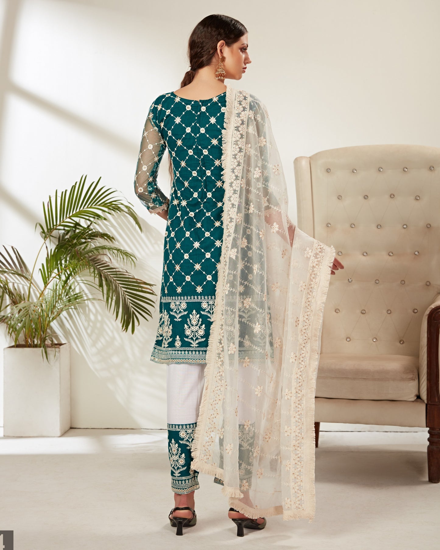 Woman in teal and white traditional outfit with a beige chair and plant in the background