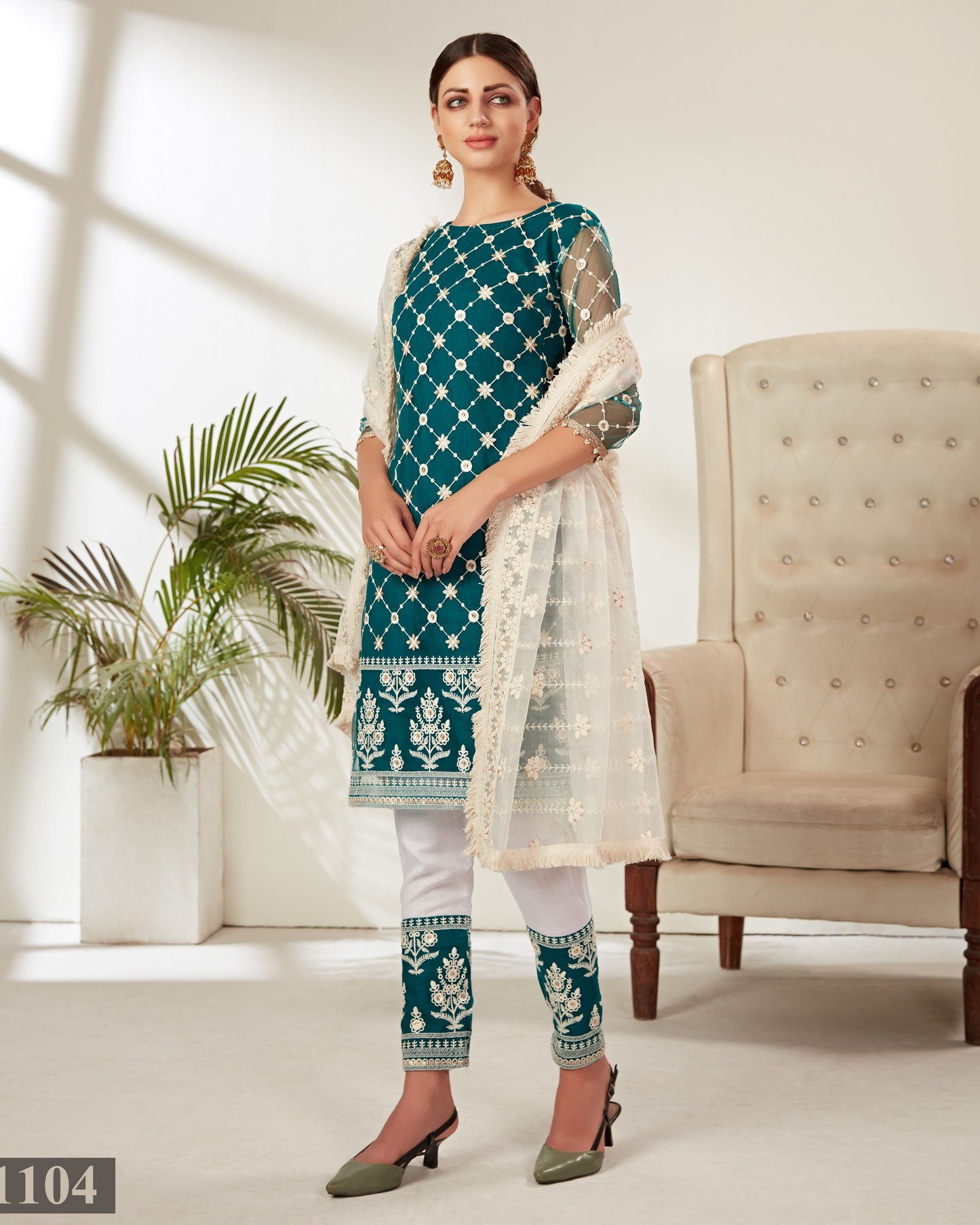 Woman wearing a teal and white traditional outfit in a bright room with a plant and chair.
