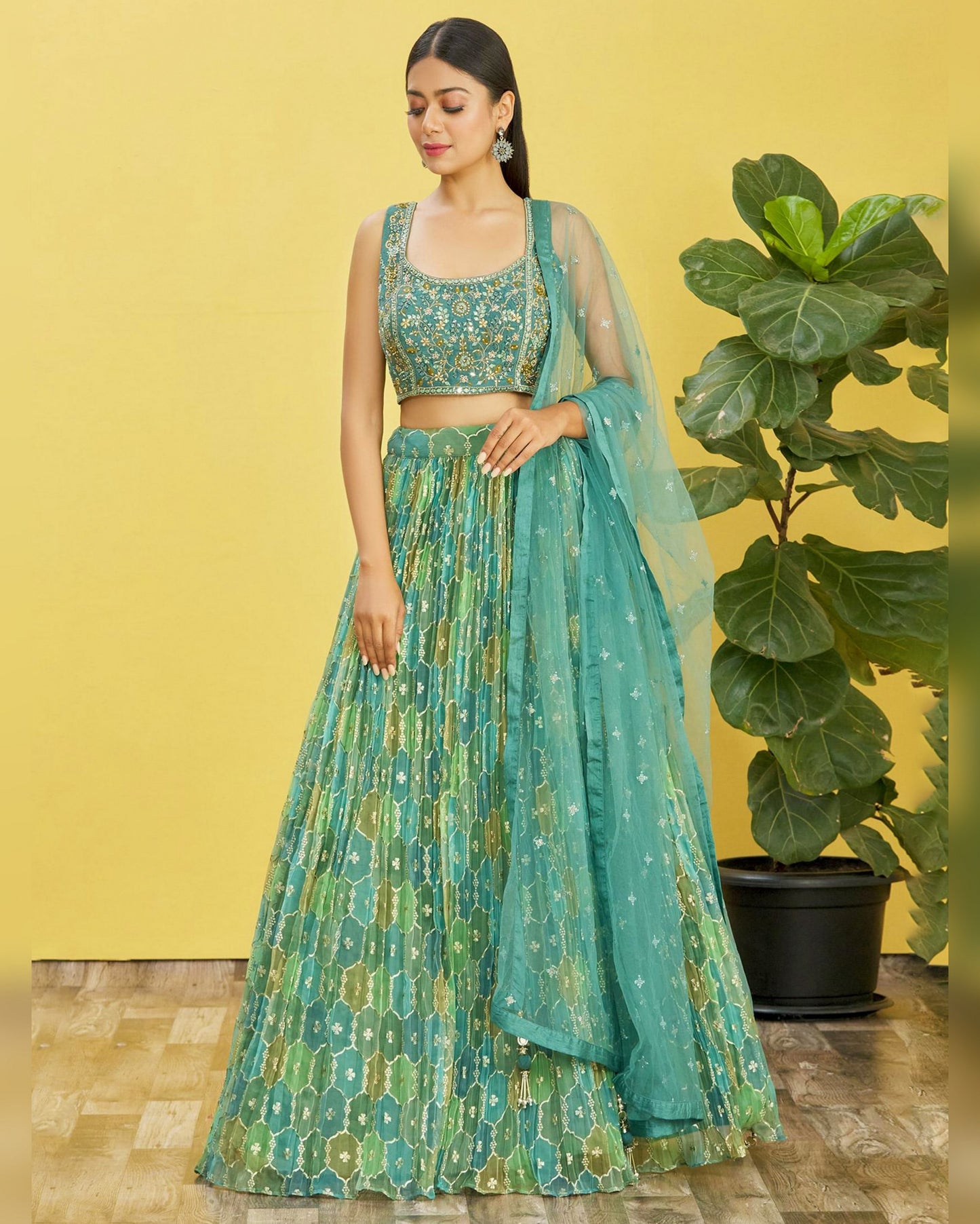 Woman in a green and blue patterned outfit standing against a yellow wall with a plant.